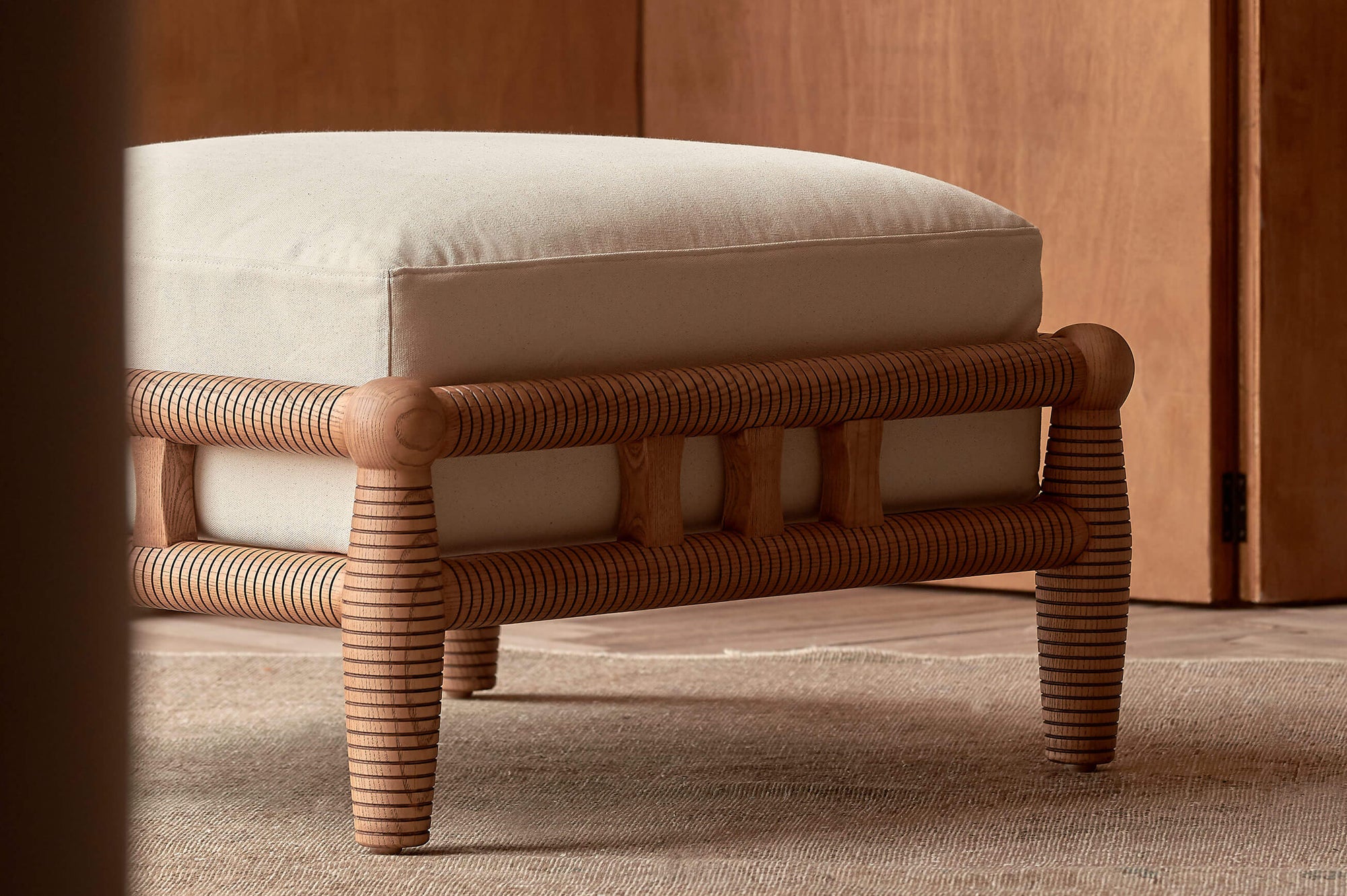 Gio Ottoman in a Heritage Ash wood frame with Cotton Canvas Beach Walk slipcover, placed on top of a rug