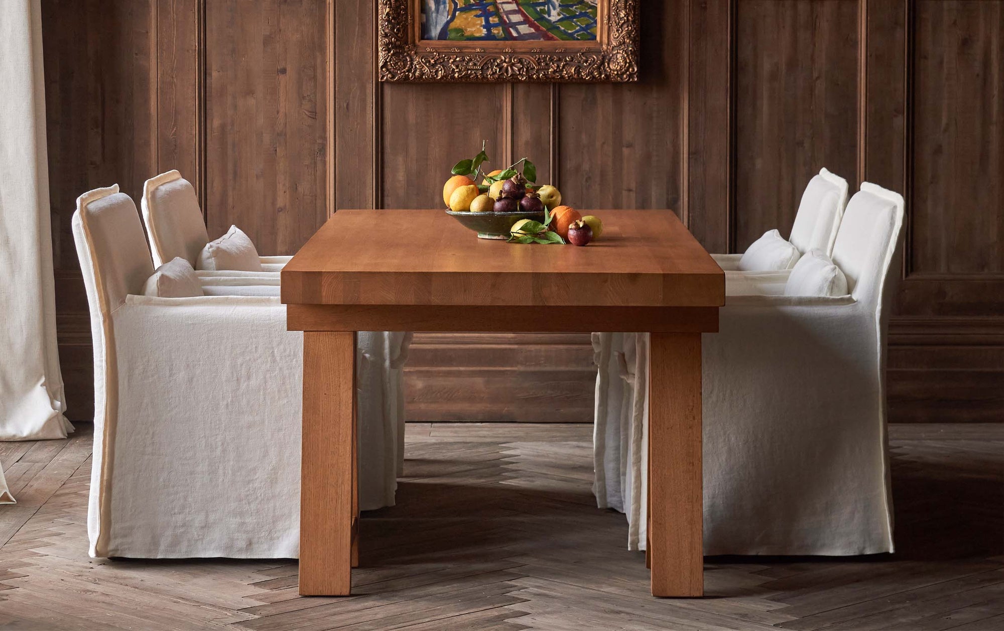 Two sets of Melo Dining Chairs in Water Lily, a white Light Weight Linen with warm undertones, placed around a Rylance Dining Table in a wood paneled room. The table is decorated with a fruit bowl centerpiece.