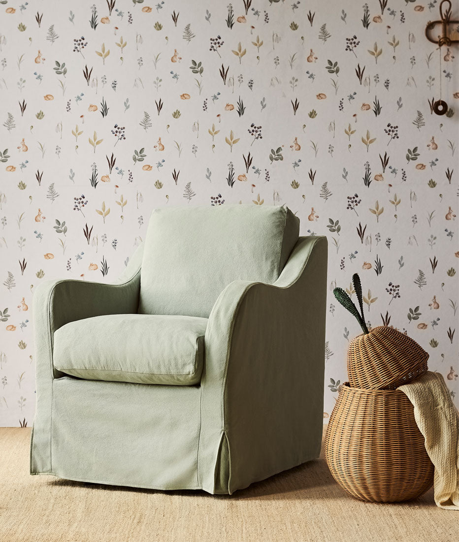 Esme Glider Chair in Thread Dyed Cotton Linen Hello Aloe placed in front of a wall covered in floral wallpaper