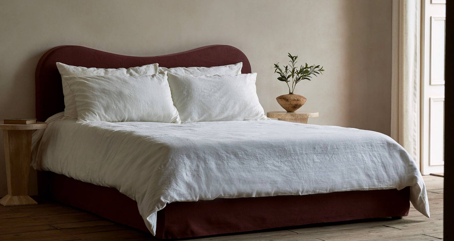 An Esmé Bed in Thread Dyed Cotton Linen Summer Plum with white pillows and bedding placed between two side tables.