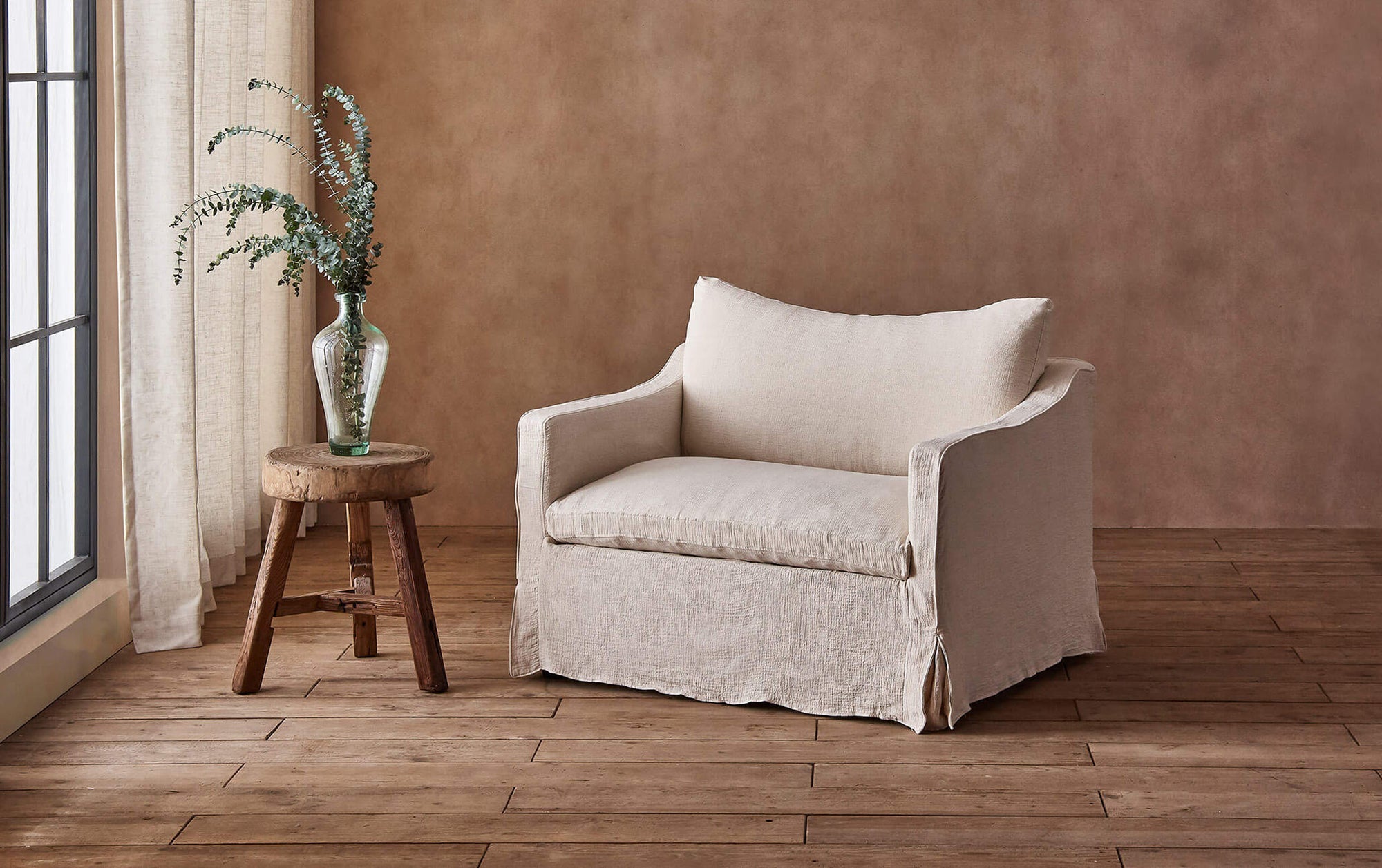 Amelia Chair in Corn Silk, a light beige Washed Cotton Linen, placed in a sunlit room next to a vase of flowers on top of a stool