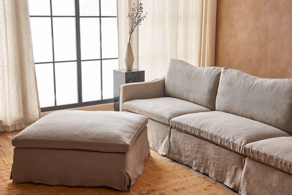 Close up of Amelia 5-piece Chaise Sectional Sofa in Jasmine Rice, a light warm greige Medium Weight Linen, infront of a bright window and a stone pedestal holding a vase of flowers