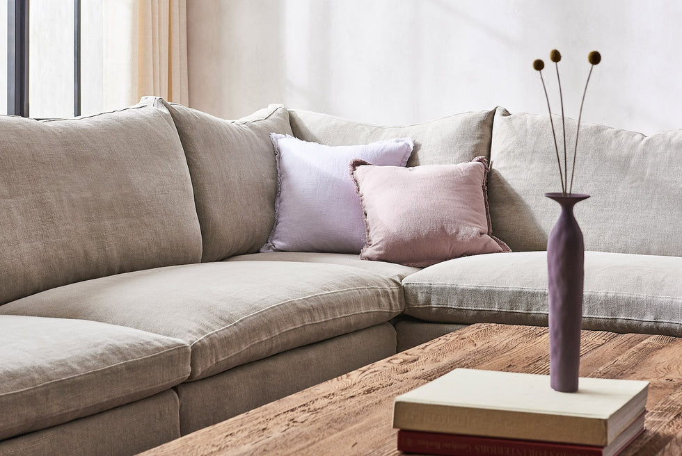 Close up of Amelia Corner Sectional Sofa in Jasmine Rice, a light warm greige Medium Weight Linen, placed around a Kai Coffee Table holding books and thing purple vase