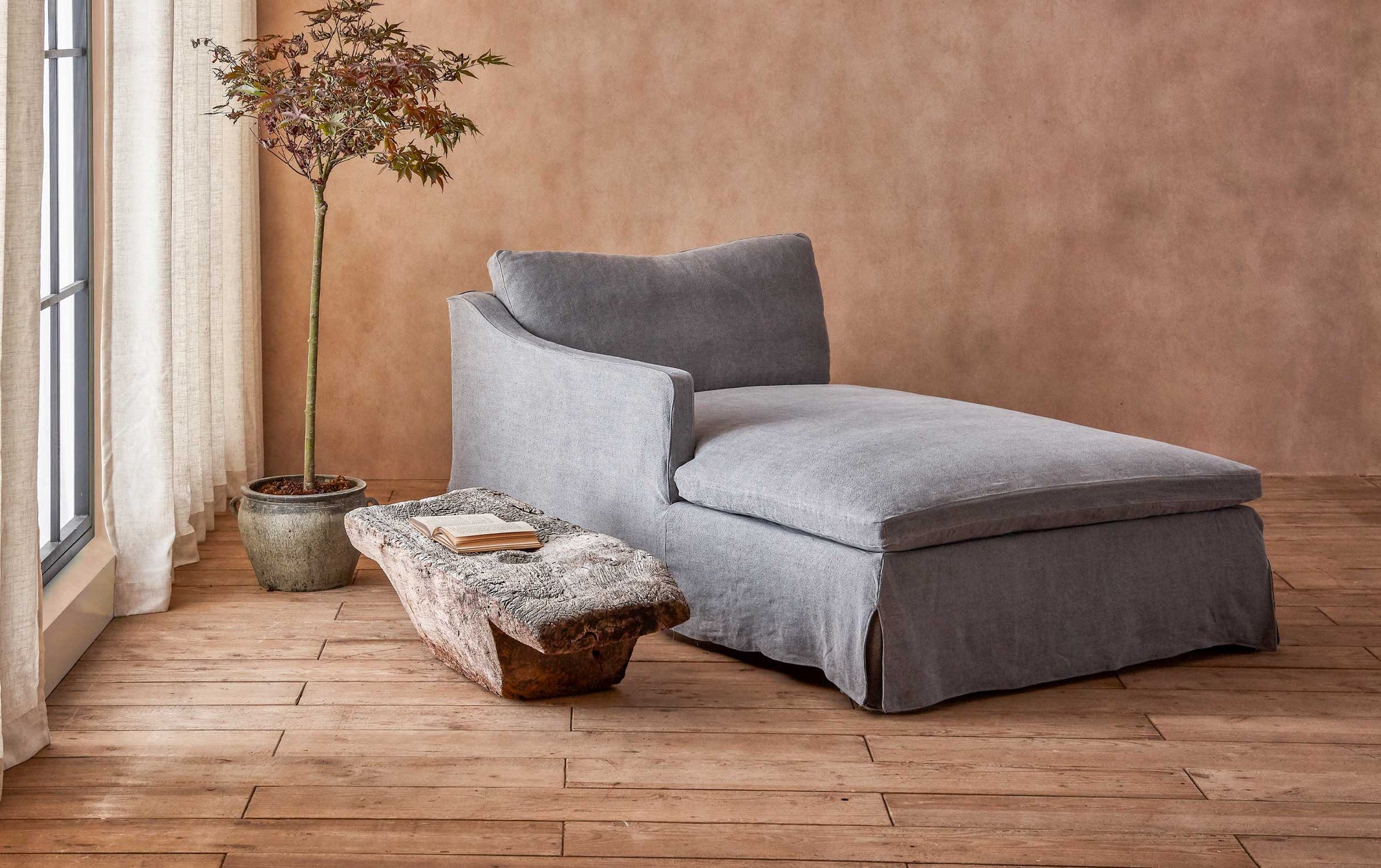 Amelia Left Arm Facing Daybed in Ink Cap, a medium cool grey Light Weight Linen, placed in a warmly lit room next to a stone table and a potted tree