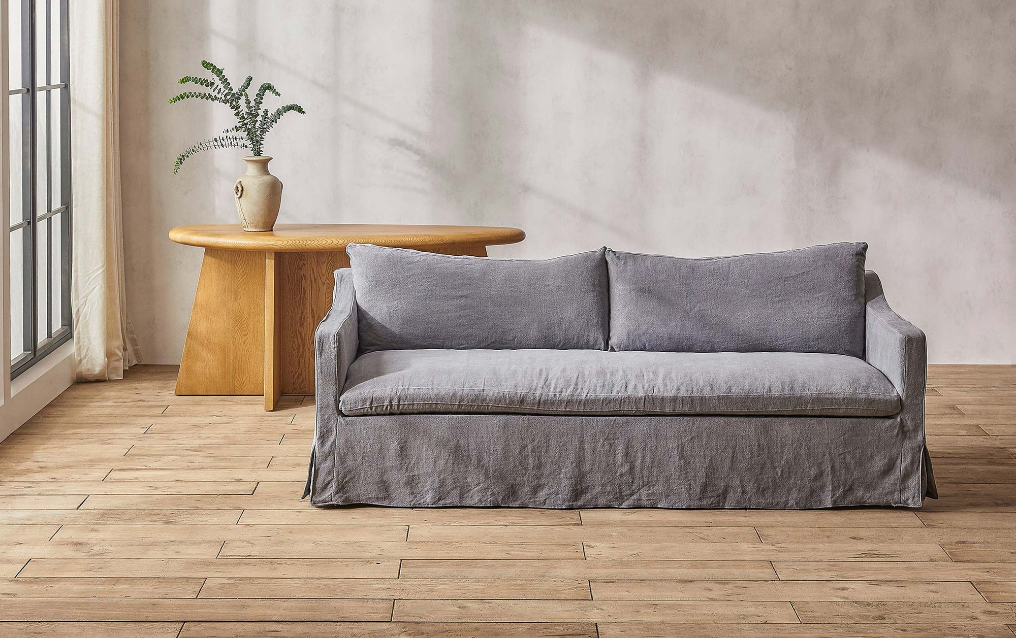 Amelia sofa in gray light weight linen placed in a sunlit room in front of a vase of ferns on a table