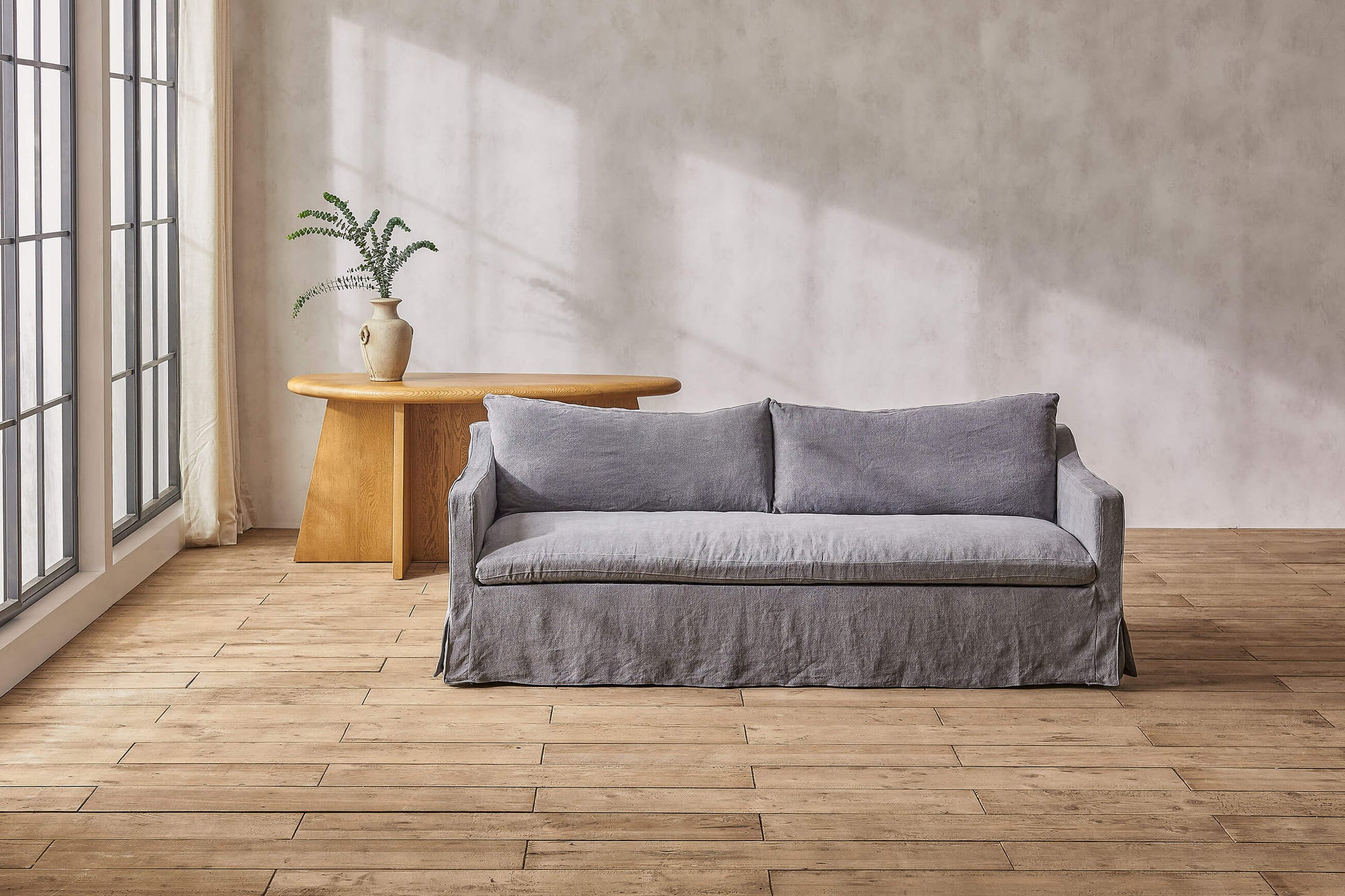 Amelia sofa in gray light weight linen placed in a sunlit room in front of a vase of ferns on a table