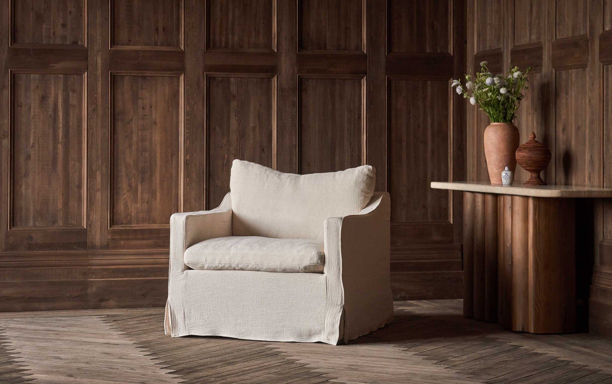Amelia Swivel Chair in Corn Silk, a light beige Washed Cotton Linen, placed in a wood panneled room next to an Enzo Console Table decorated with a vase of flowers