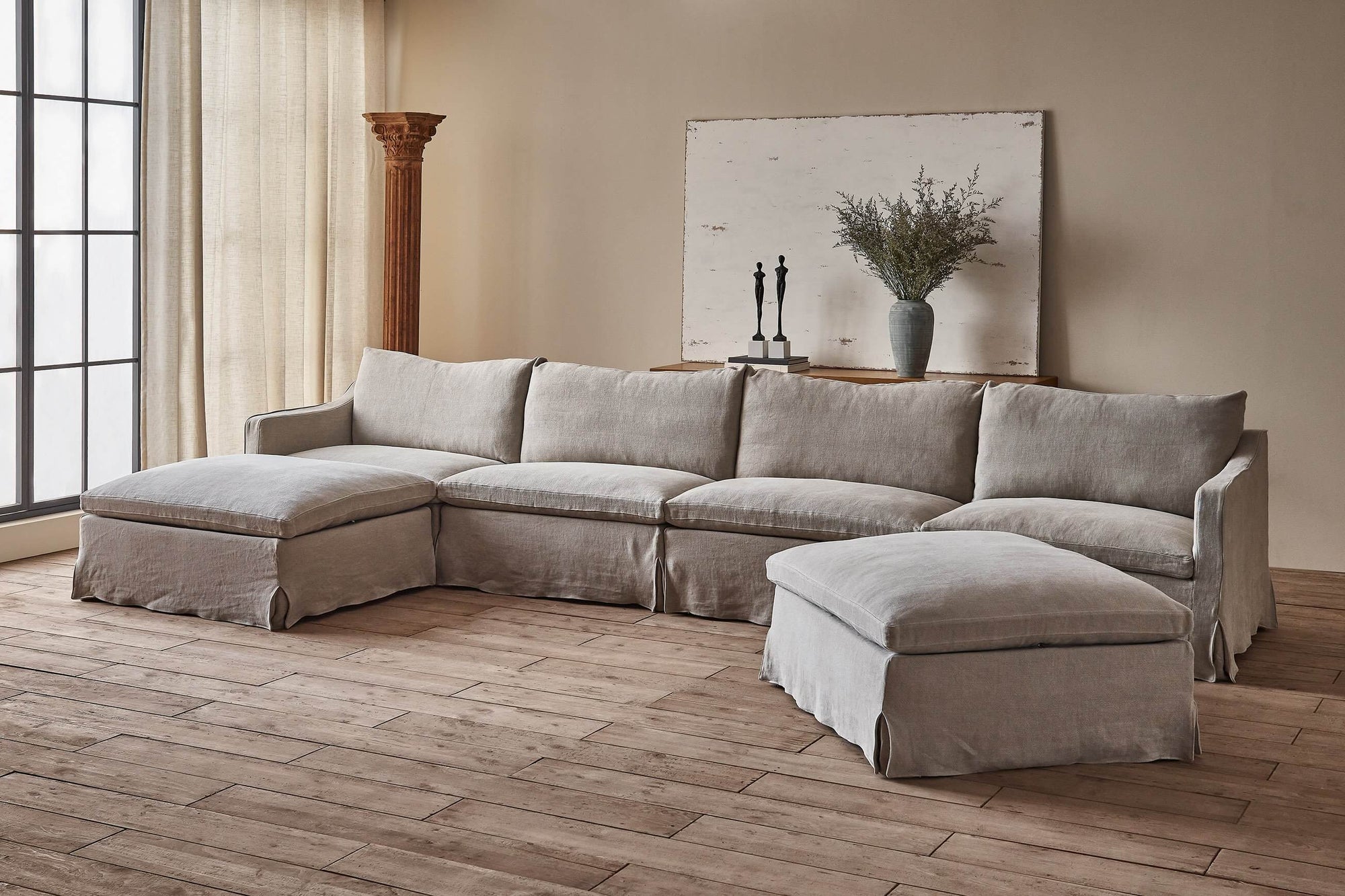 Amelia U-Shape Sectional Sofa in Jasmine Rice, a light warm greige Medium Weight Linen, placed in a bright room with a wooden pillar and a console table decorated with two small statues, wildflowers in a vase, and a white wooden painting.