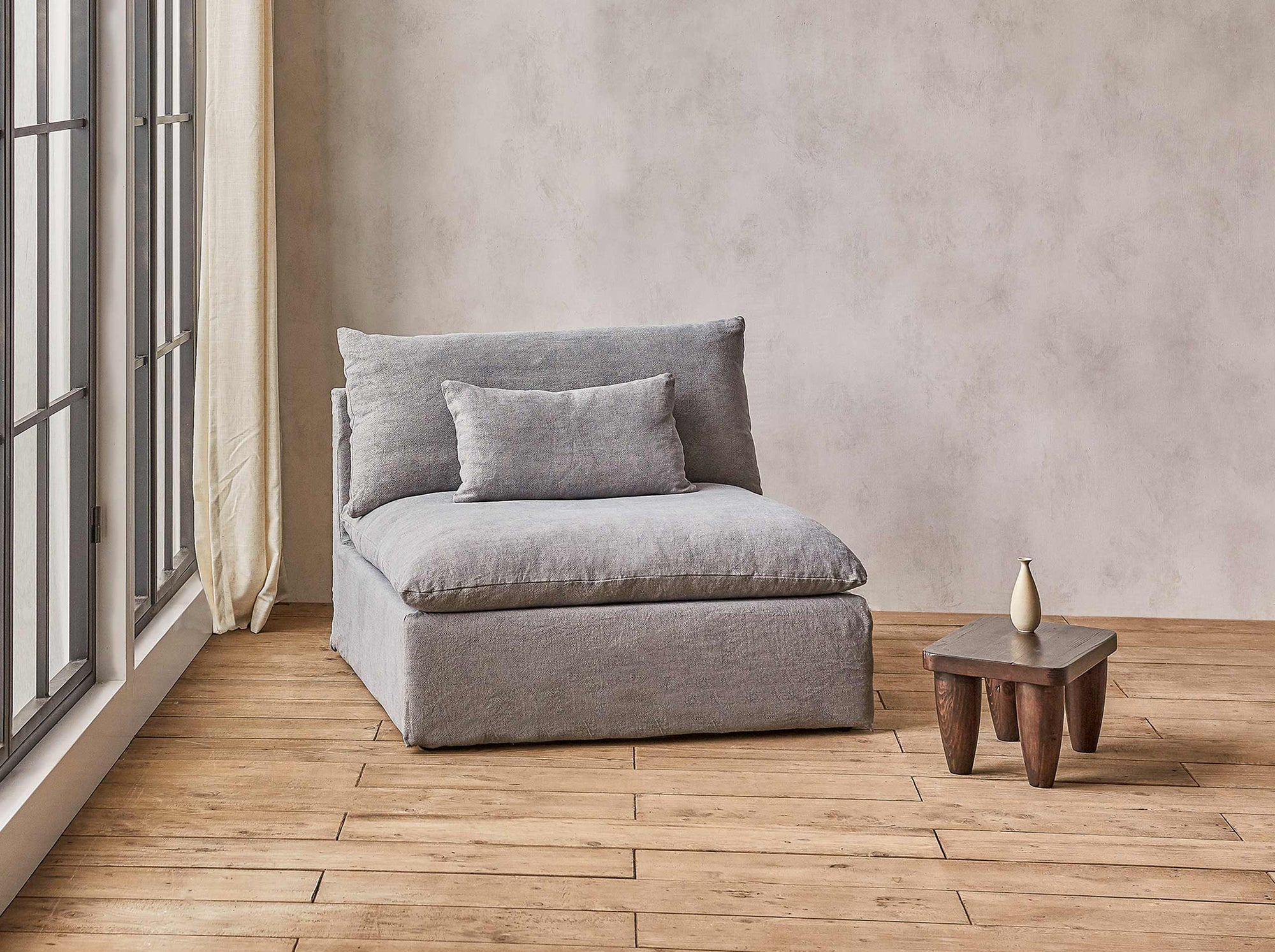 Aria Chair in Ink Cap, a medium cool grey Light Weight Linen, placed in a sunlit room next to a window and a side table