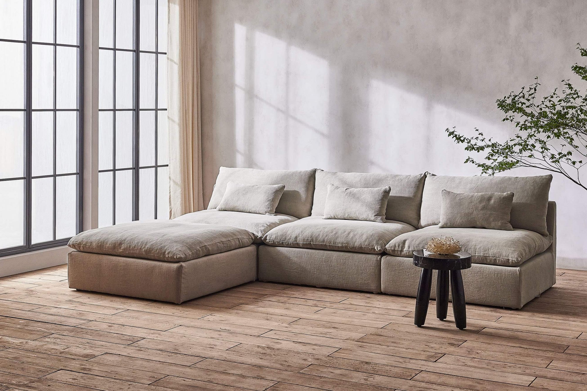 Aria 4-piece Chaise Sectional Sofa in Jasmine Rice, a light warm greige Medium Weight Linen, placed in a sunlit room with a large windown and a black stone stool