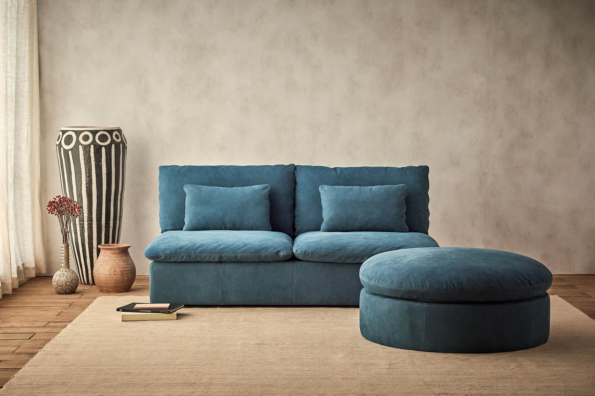 Aria 72" Leather Sofa in North Atlantic, a dark teal blue Meridian Leather, with a matching round ottoman, next to a few decorative vases