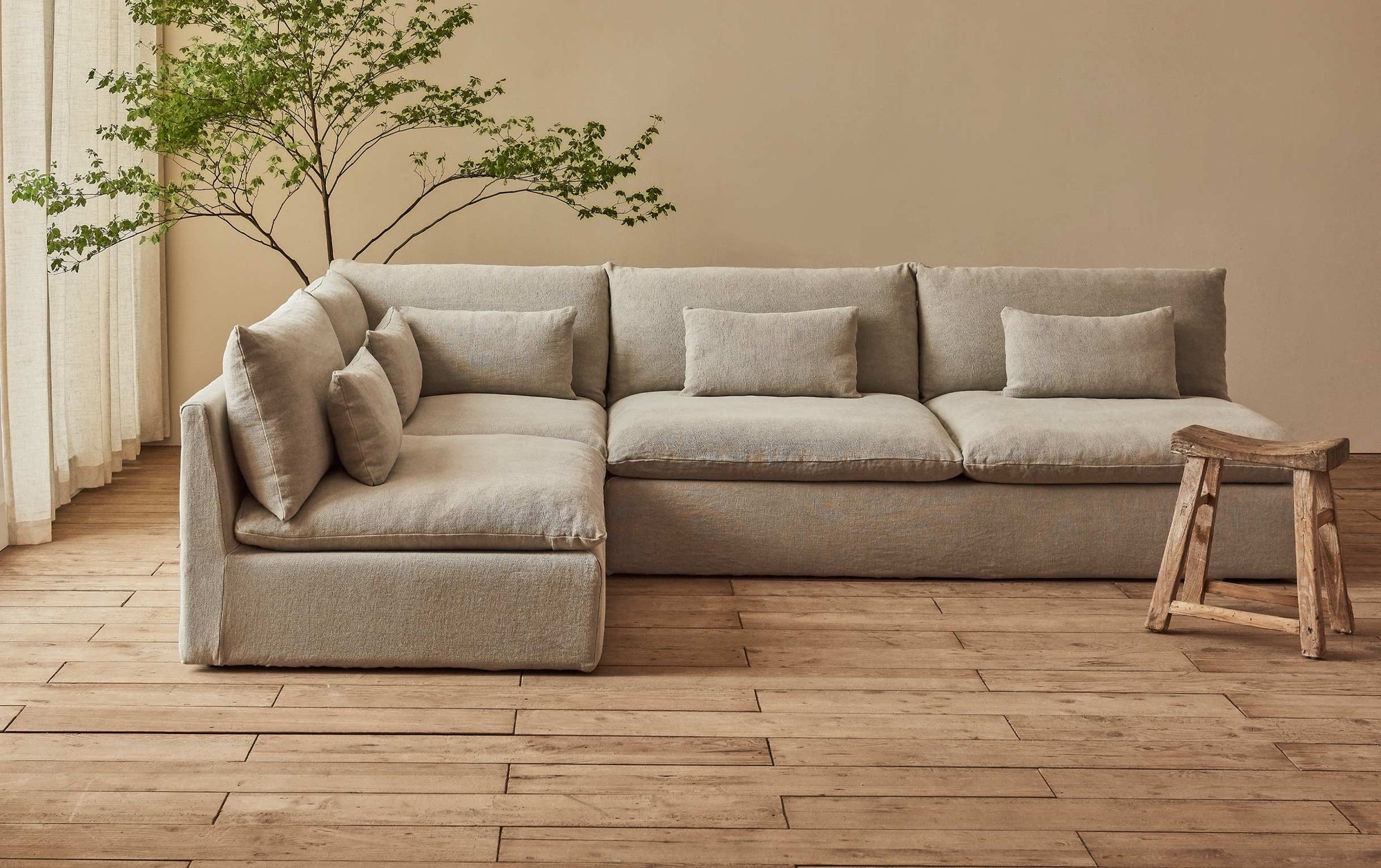 Aria Long L-Shape Sectional Sofa in Jasmine Rice, a light warm greige Medium Weight Linen, placed in a sunlit room between a potted tree and a stool