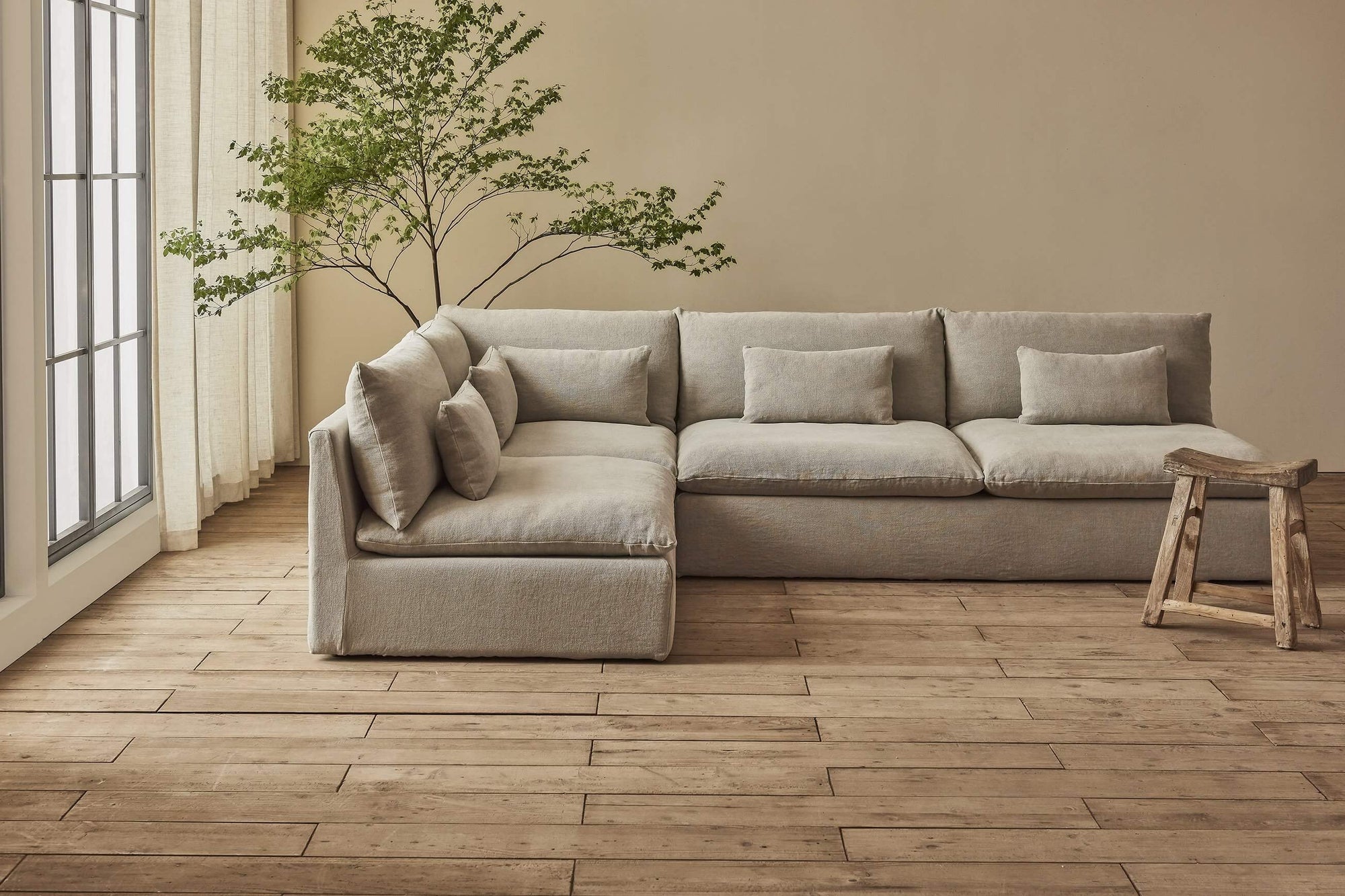 Aria Long L-Shape Sectional Sofa in Jasmine Rice, a light warm greige Medium Weight Linen, placed in a sunlit room with a wooden stool and a large plant