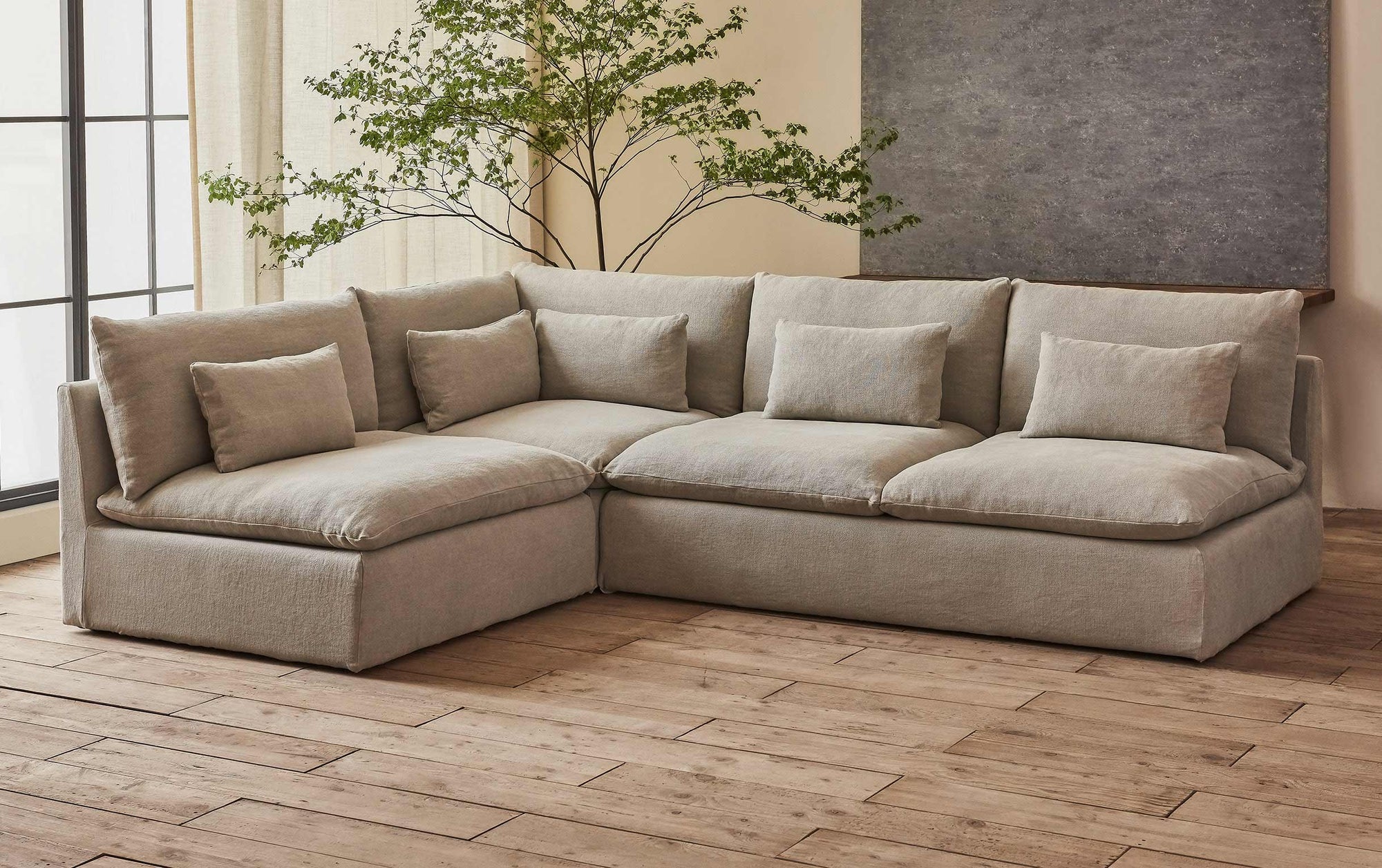 Aria Short L-Shape Sectional Sofa in Jasmine Rice, a light warm greige Medium Weight Linen, placed in a sunlit room in front of a potted tree