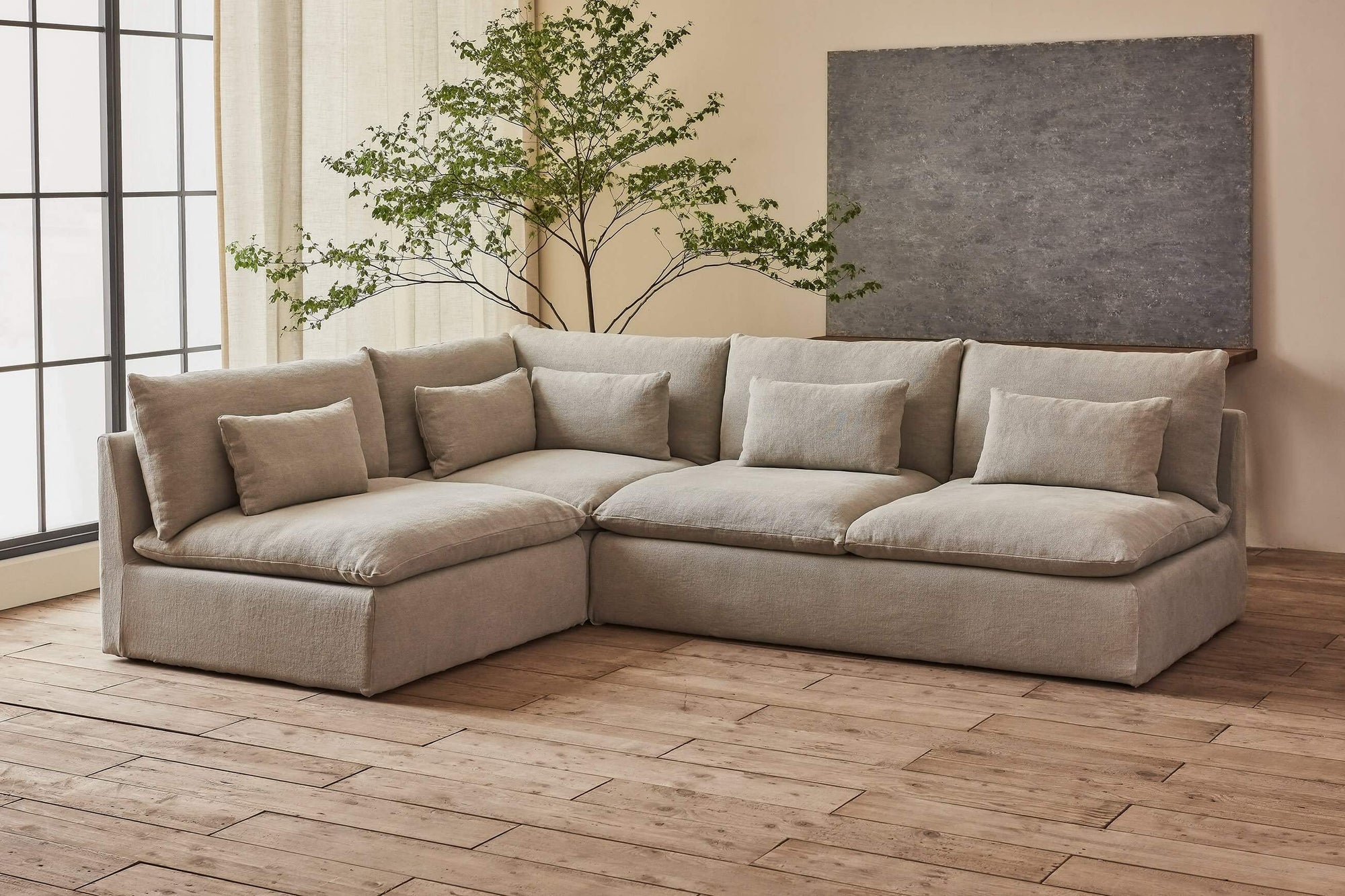 Aria Short L-Shape Sectional Sofa in Jasmine Rice, a light warm greige Medium Weight Linen, placed in a sunlit room with a large window and a stone wall hanging