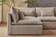 Slide #6 Close up of Aria Short L-Shape Sectional Sofa in Jasmine Rice, a light warm greige Medium Weight Linen, placed in a sunlit room in front of a tall plant and a stone wall hanging