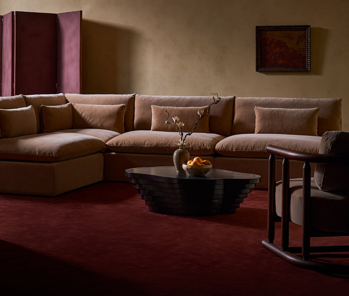 A styled living room scene featuring a Sixpenny Aria L-shaped washed cotton velvet sectional sofa with matching cushions, a Sixpenny Terra coffee table, a Sixpenny Gio chair and a deep burgundy carpet against a warm beige wall.