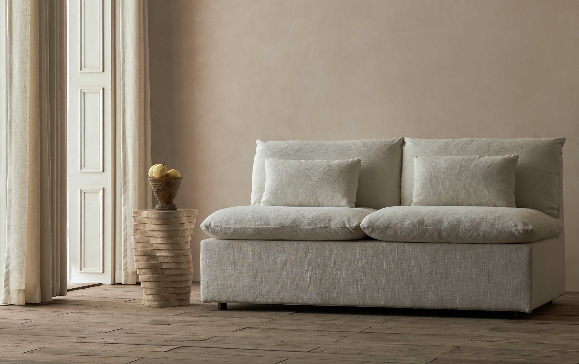 Aria Sleeper Sofa in Young Coconut, a powdery white Recycled Poly Linen in a beige room with a sculpted stone side table decorated with a bowl of pears.