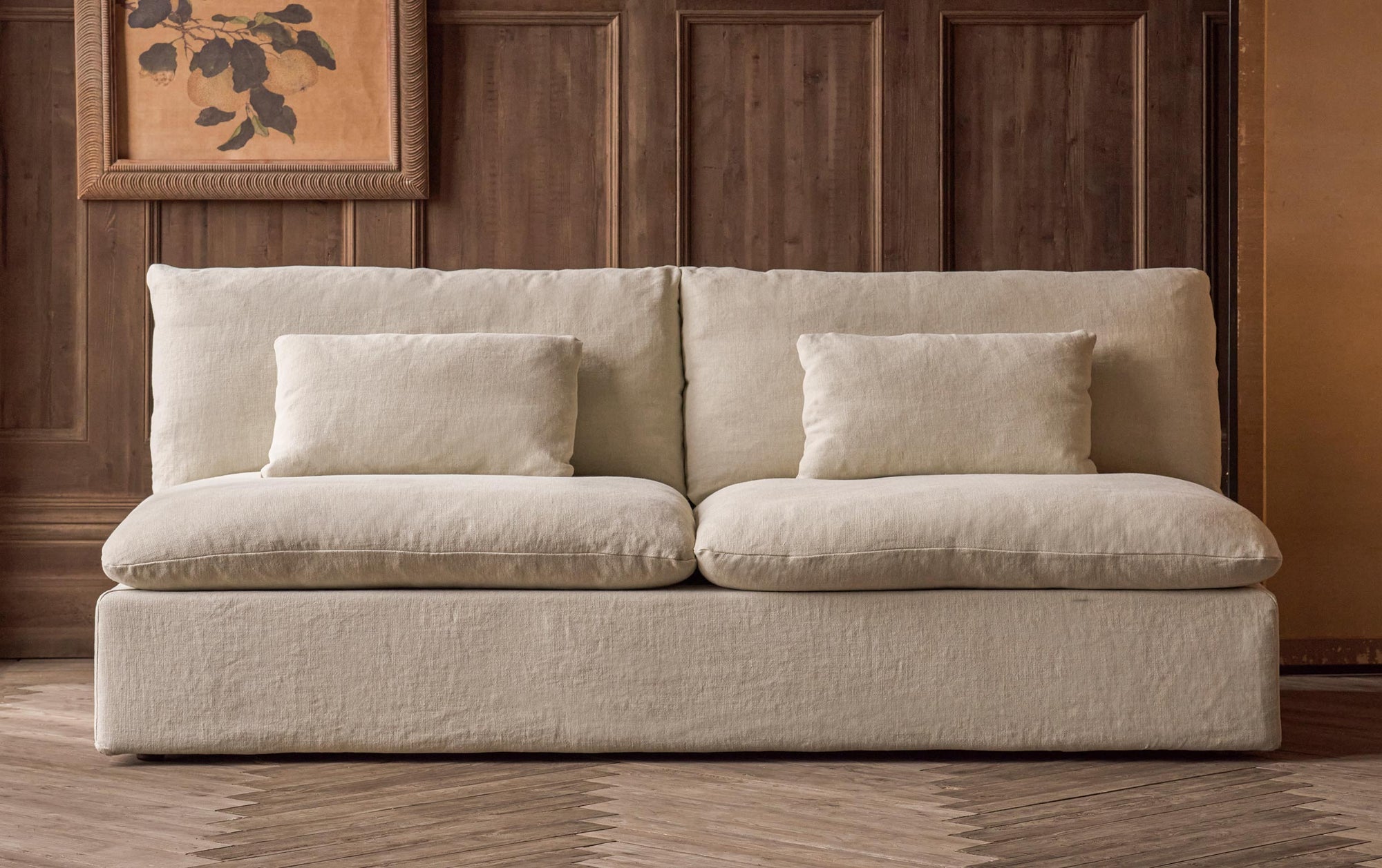 Aria 84" Sofa in Warm Oatmeal, a light warm beige Medium Weight Linen placed in front of a painting hung on a wood paneled wall. 