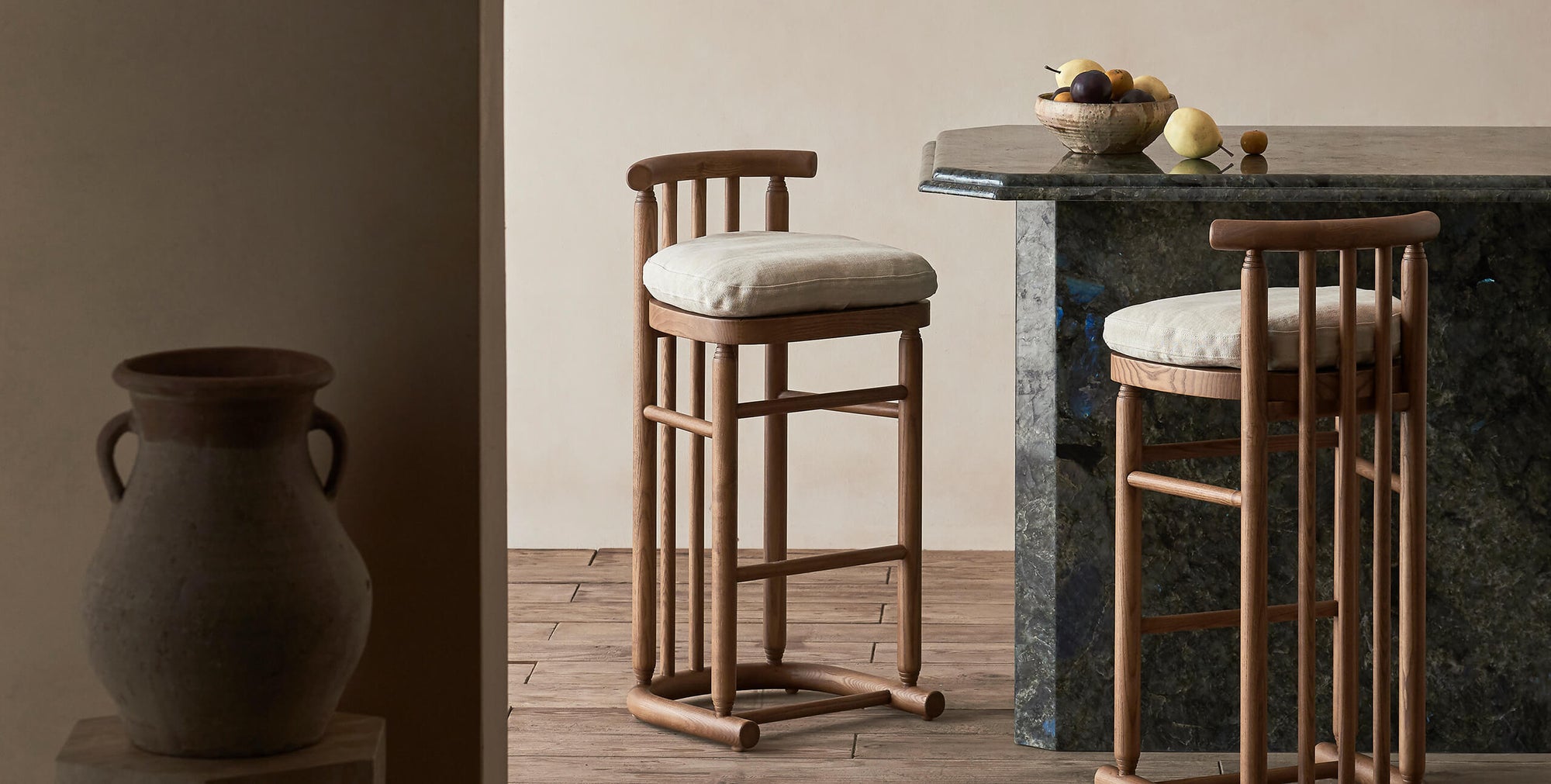 Two Bene Counter Stools in Heritage Ash wood and cushions in Warm Oatmeal, a light warm beige Medium Weight Linen, sitting at a dark stone counter holding a bowl of fruit