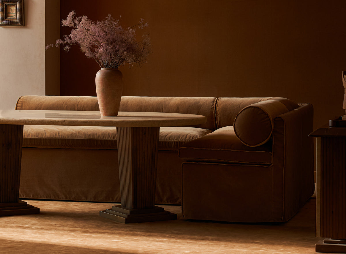 A Sixpenny Camino Banquette in Sugar Rush with a round back cushion, shown in a dimly lit living room setting with a matching Cordelia Dining table and a vase of dried flowers.