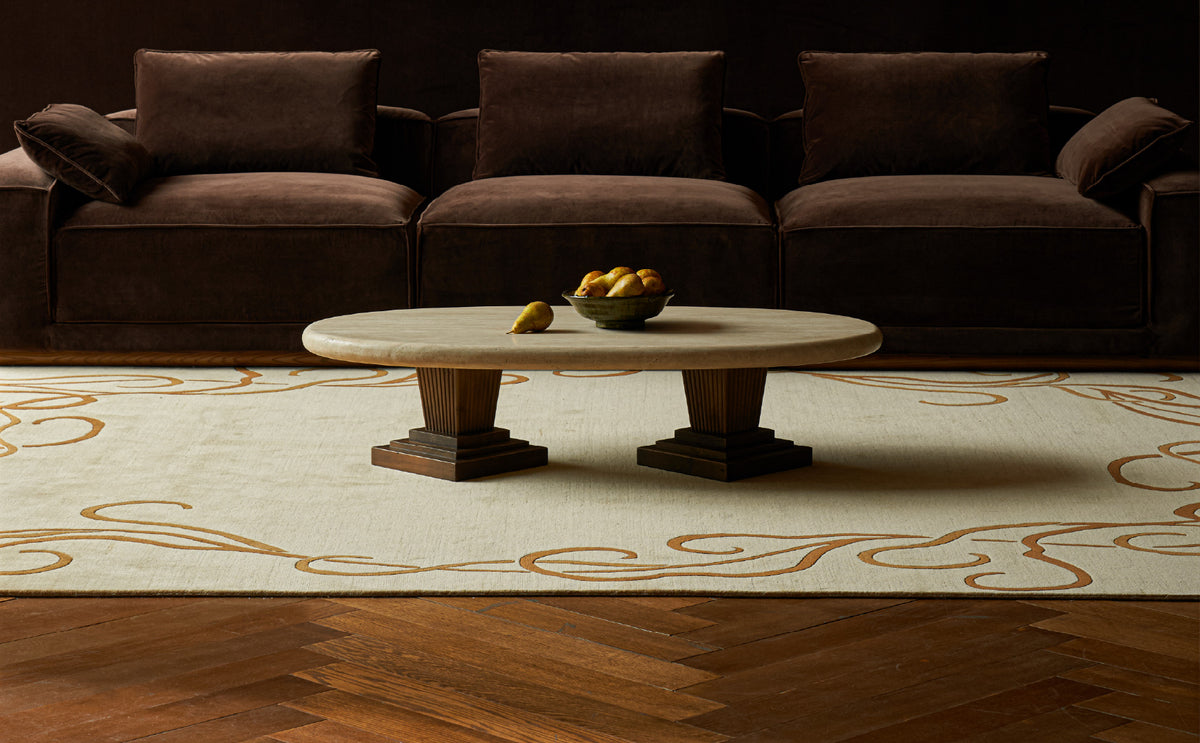 A Sixpenny Cordelia Galata travertine coffee table with a light stone top and dark wooden pedestal legs sits on a Sixpenny Wander Rug in front of a dark brown velvet sofa.