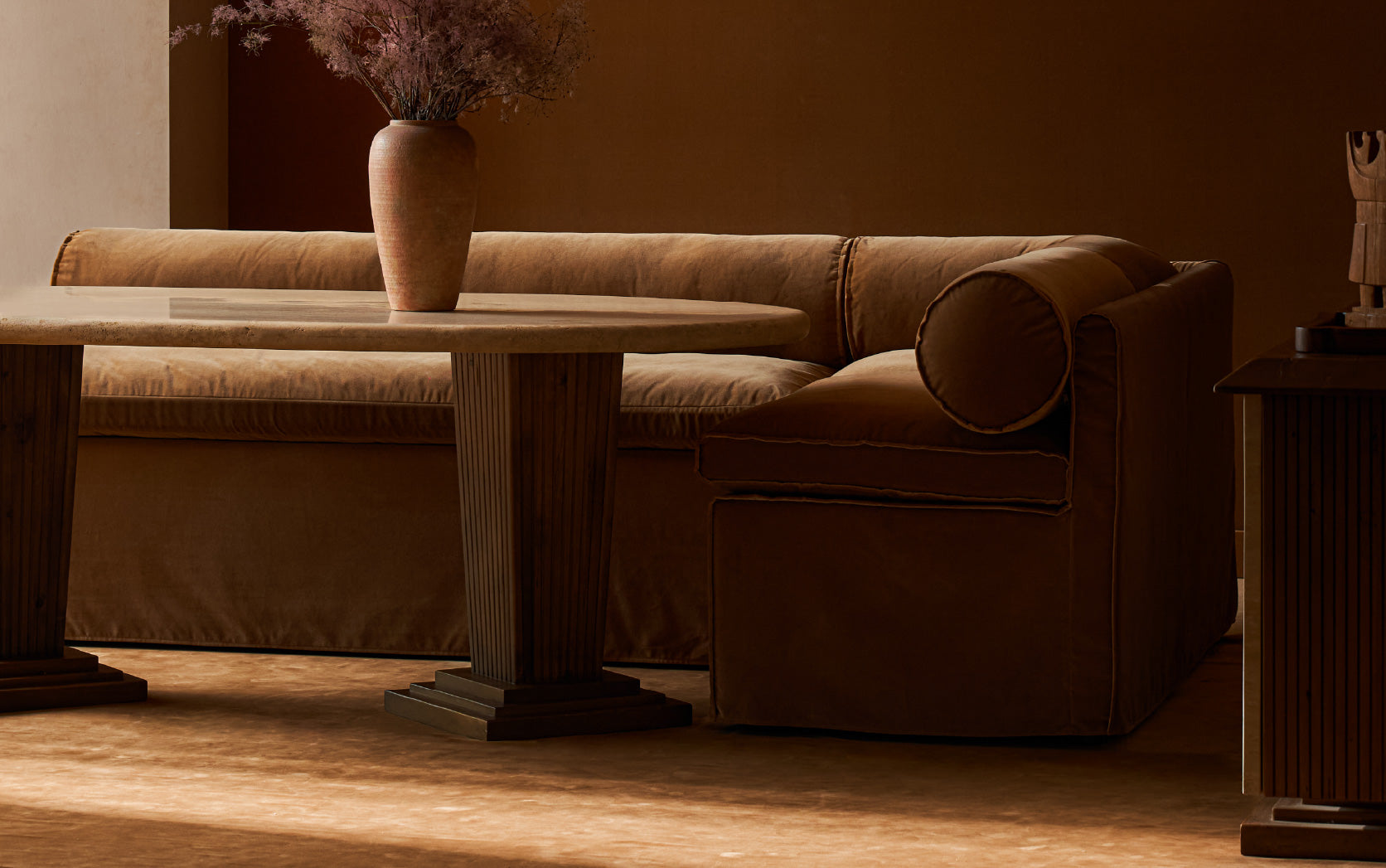 A Sixpenny Camino Banquette in Sugar Rush with a round back cushion, shown in a dimly lit living room setting with a matching Cordelia Dining table and a vase of dried flowers.