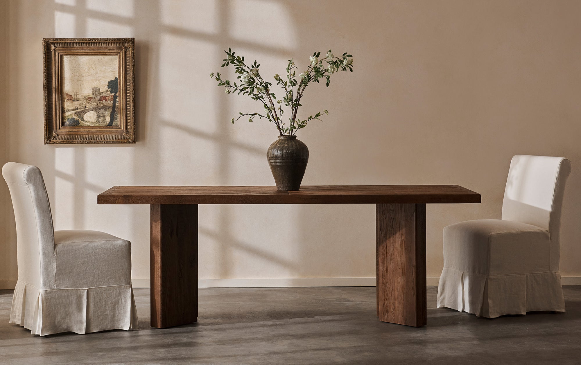 A set of Dalia Armless Dining Chairs in Water Lily, a white Light Weight Linen with warm undertones, placed at a Tango Dining Table in Heritage Oak, which is set with a vase of flowers as its centerpiece