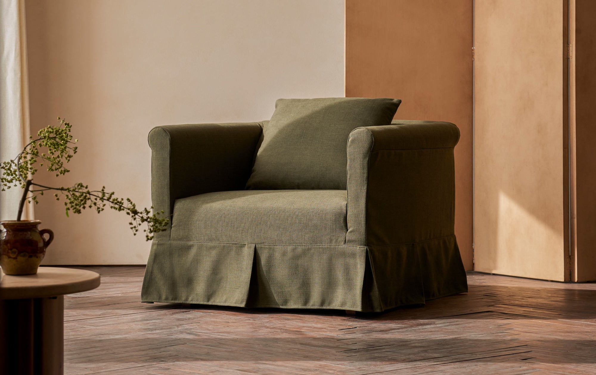 Dalia Chair in New Leaf, a forest green linen-like recycled polyester, placed in a sunny room with a decorative wooden screen and a round side table holding a potted plant.