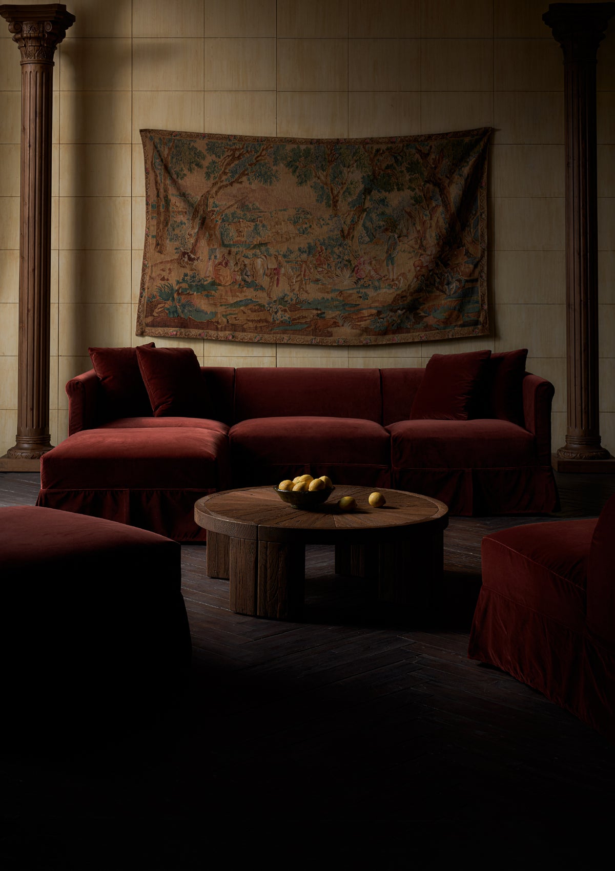 A moody, dimly lit living room featuring a Sixpenny Dalia Chaise Sectional washed cotton velvet in Bing Cherry with matching pillows, a round wooden coffee table with lemons, and a large vintage tapestry hanging on a tiled wall between two dark columns.
