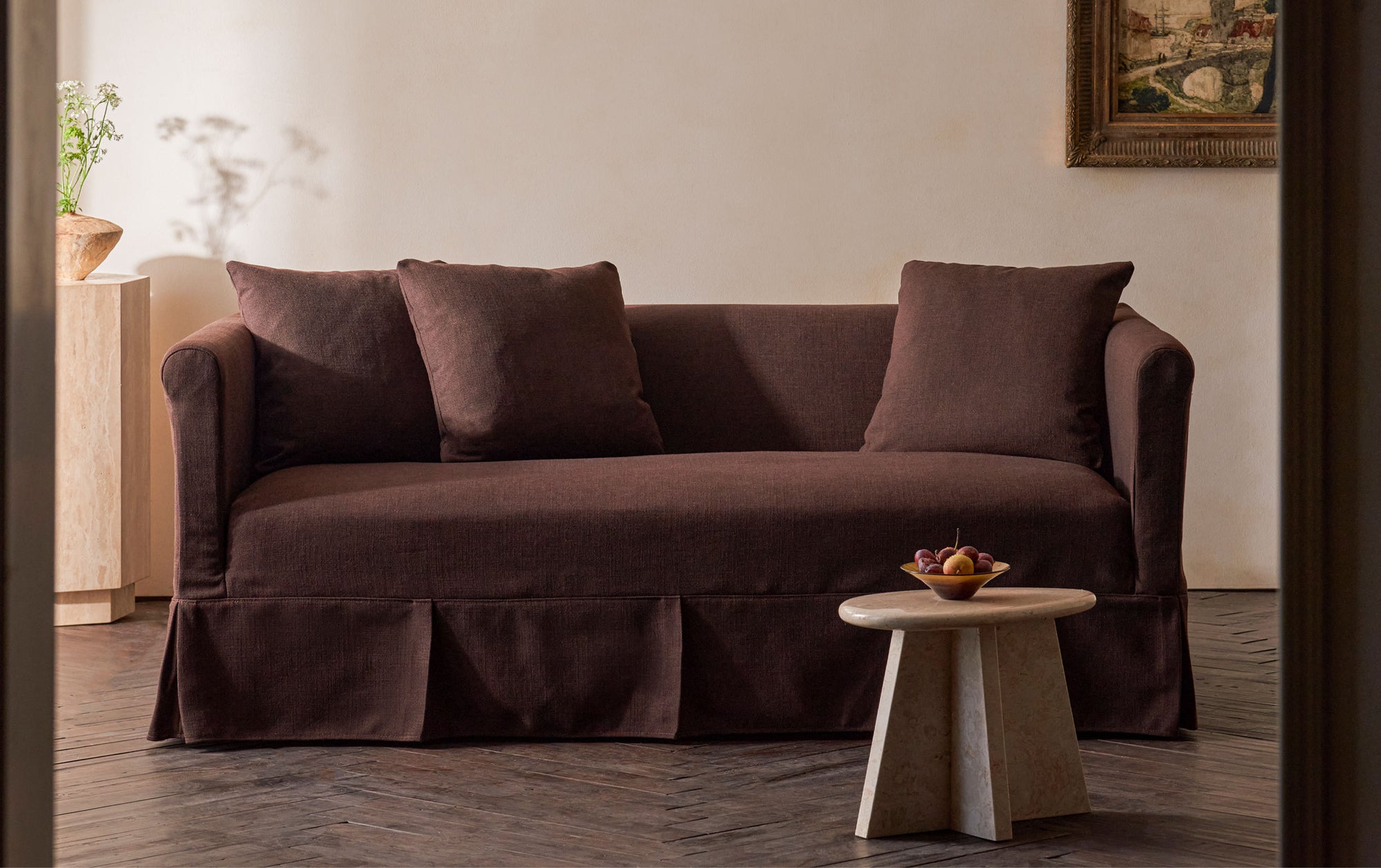 Dalia 72" Sofa in Common Ground, a chocolate brown linen-like recycled polyester, placed in a warmly lit room decorated with wall art, flower, and a small stone side table holding a bowl of fruit