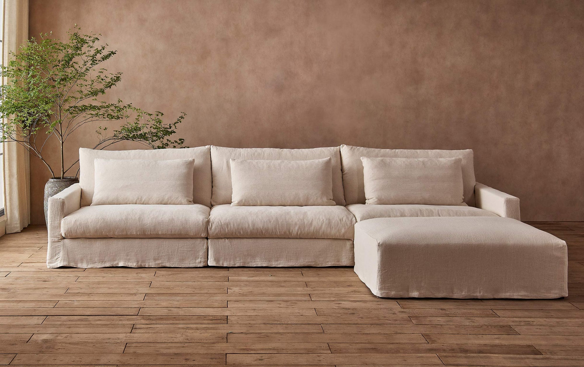 Devyn 4-piece Chaise Sectional Sofa in Corn Silk, a light beige Washed Cotton Linen, placed in front of a warm, golden-brown wall and a large potted plant