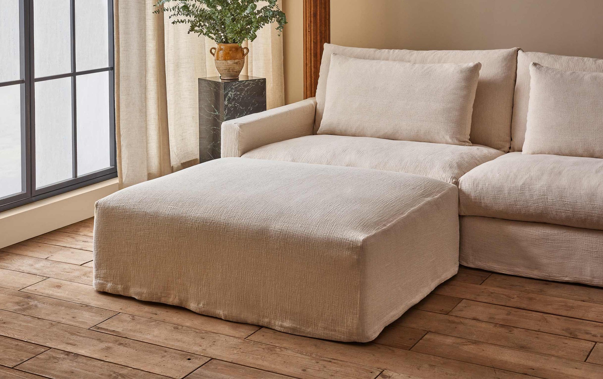 Devyn Sectional Ottoman in Corn Silk, a light beige Washed Cotton Linen, placed in front of a Devyn Sectional Sofa next to a large window and a plant on a stone pedestal