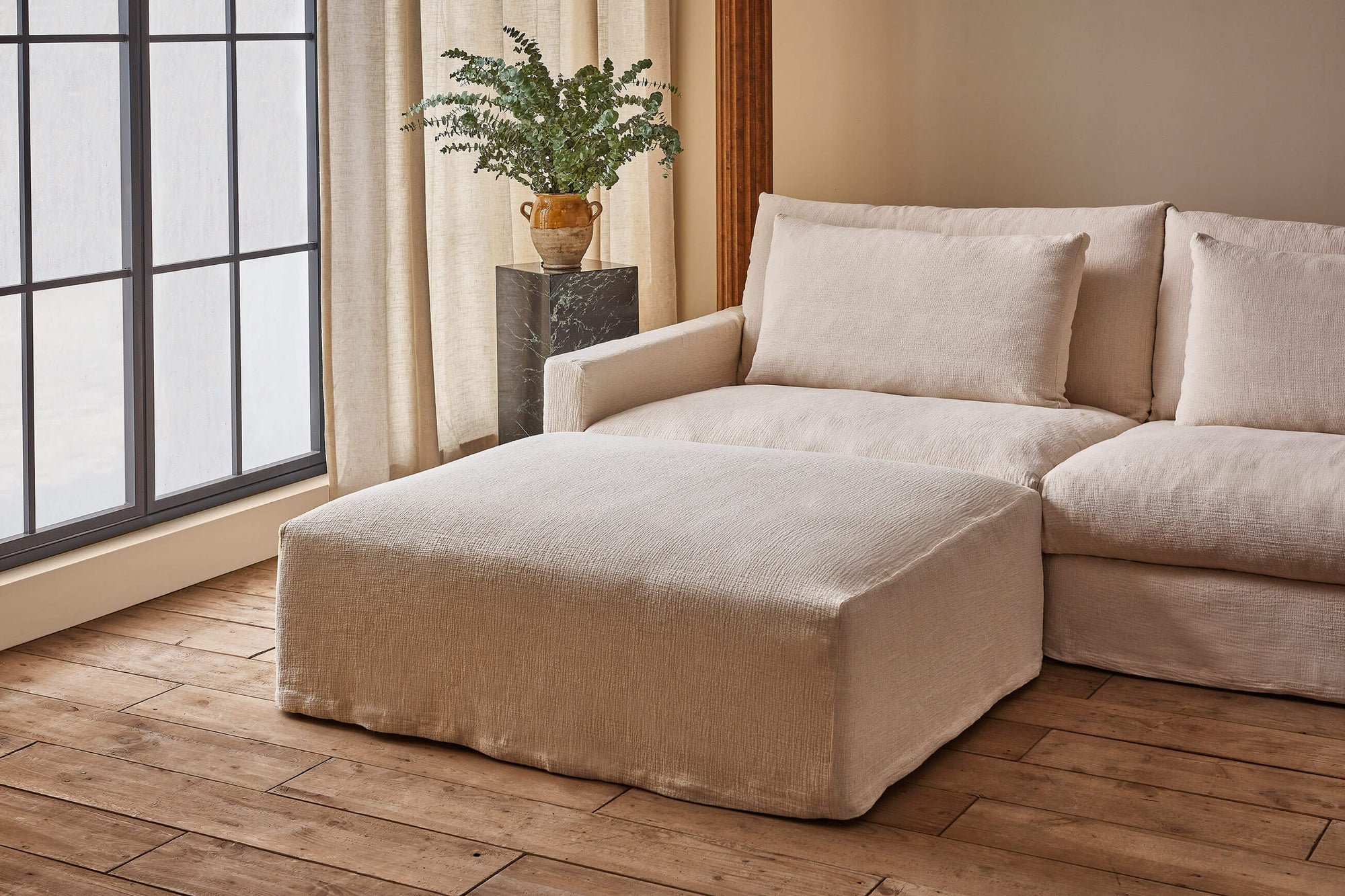 Devyn Sectional Ottoman in Corn Silk, a light beige Washed Cotton Linen, placed in front of a Devyn Sectional Sofa next to a large window and a plant on a stone pedestal