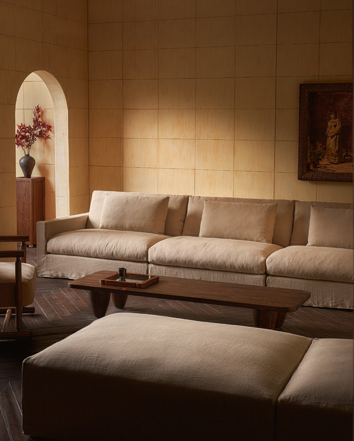 A warm, modern living room featuring a Sixpenny Devyn Sectional Sofa in a warm oatmeal matching low wooden coffee table, and a large beige ottoman in the foreground, set against a tiled beige wall.