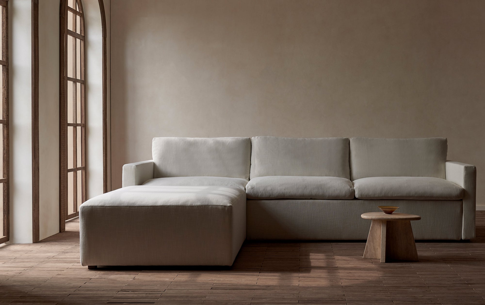 Devyn Sleeper Chaise Sectional in Young Coconut, a powdery white Recycled Poly Linen, sitting against a beige wall with a small wooden end table.