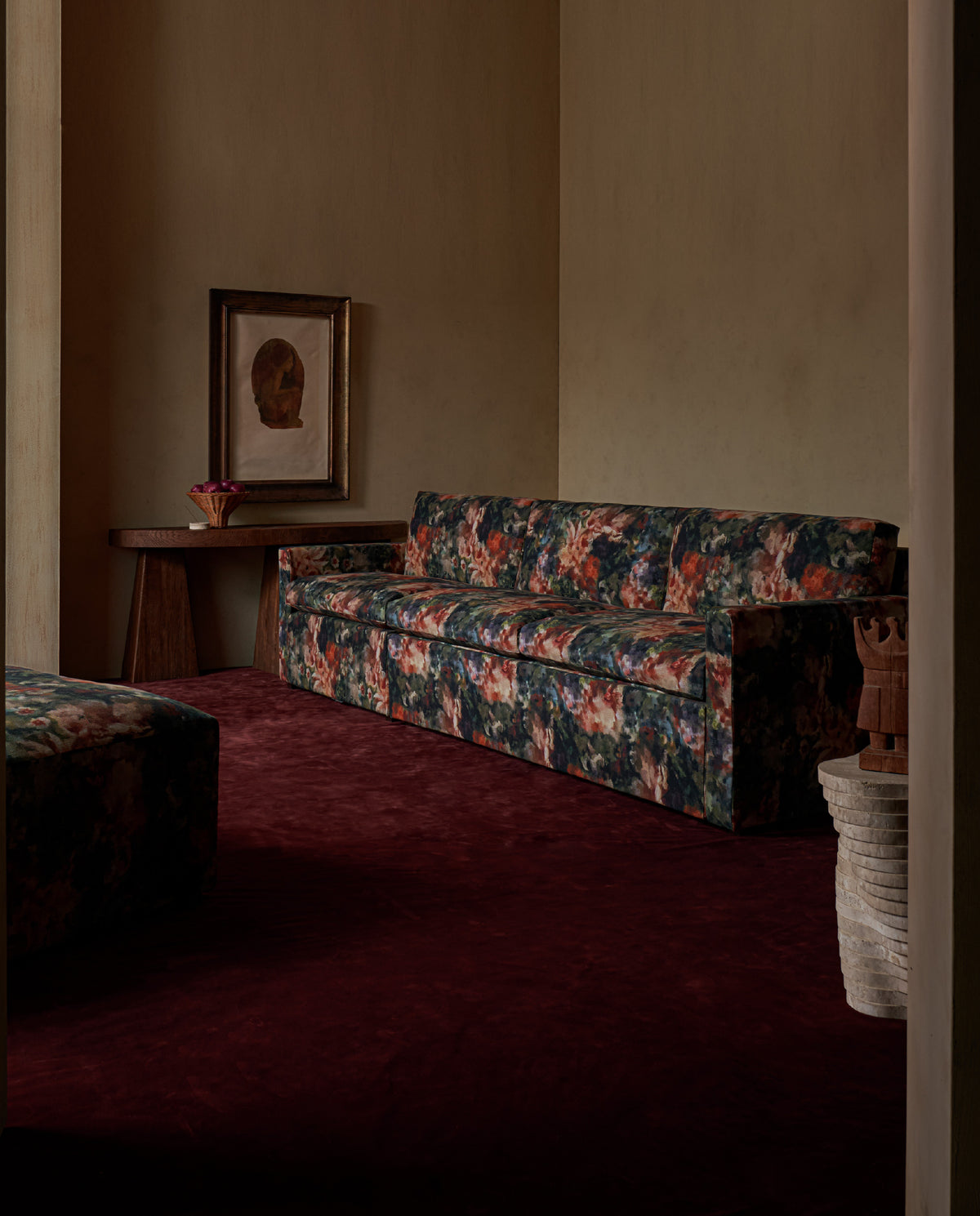 A Sixpenny Devyn sleeper sofa in wild air, a moody living room with a dark red carpet, a side table, framed wall art, and a matching ottoman partially visible in the foreground.