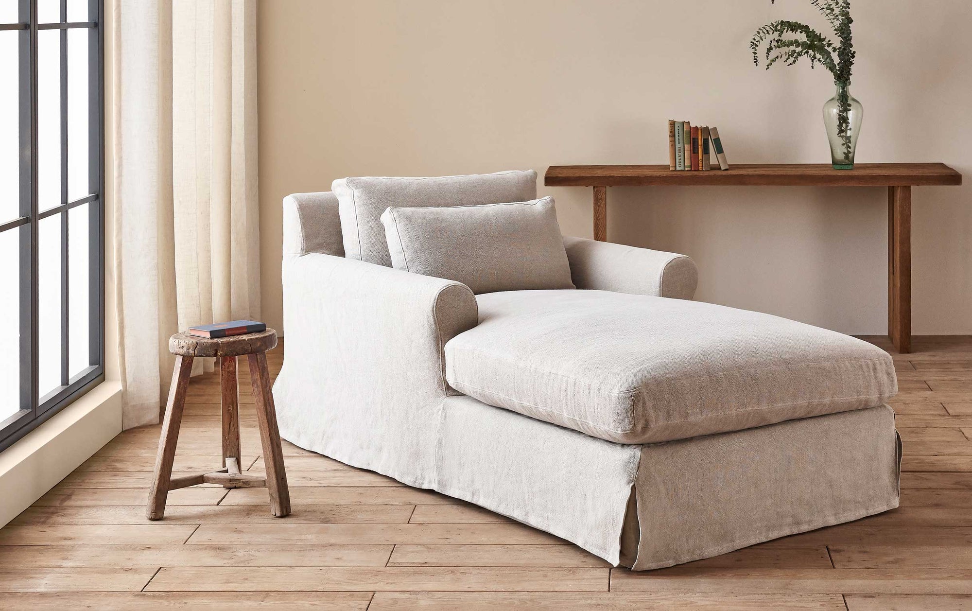 Elias Daybed in Jasmine Rice, a light warm greige Medium Weight Linen, placed in a sunlit room with a wooden stool and console table decorated with books