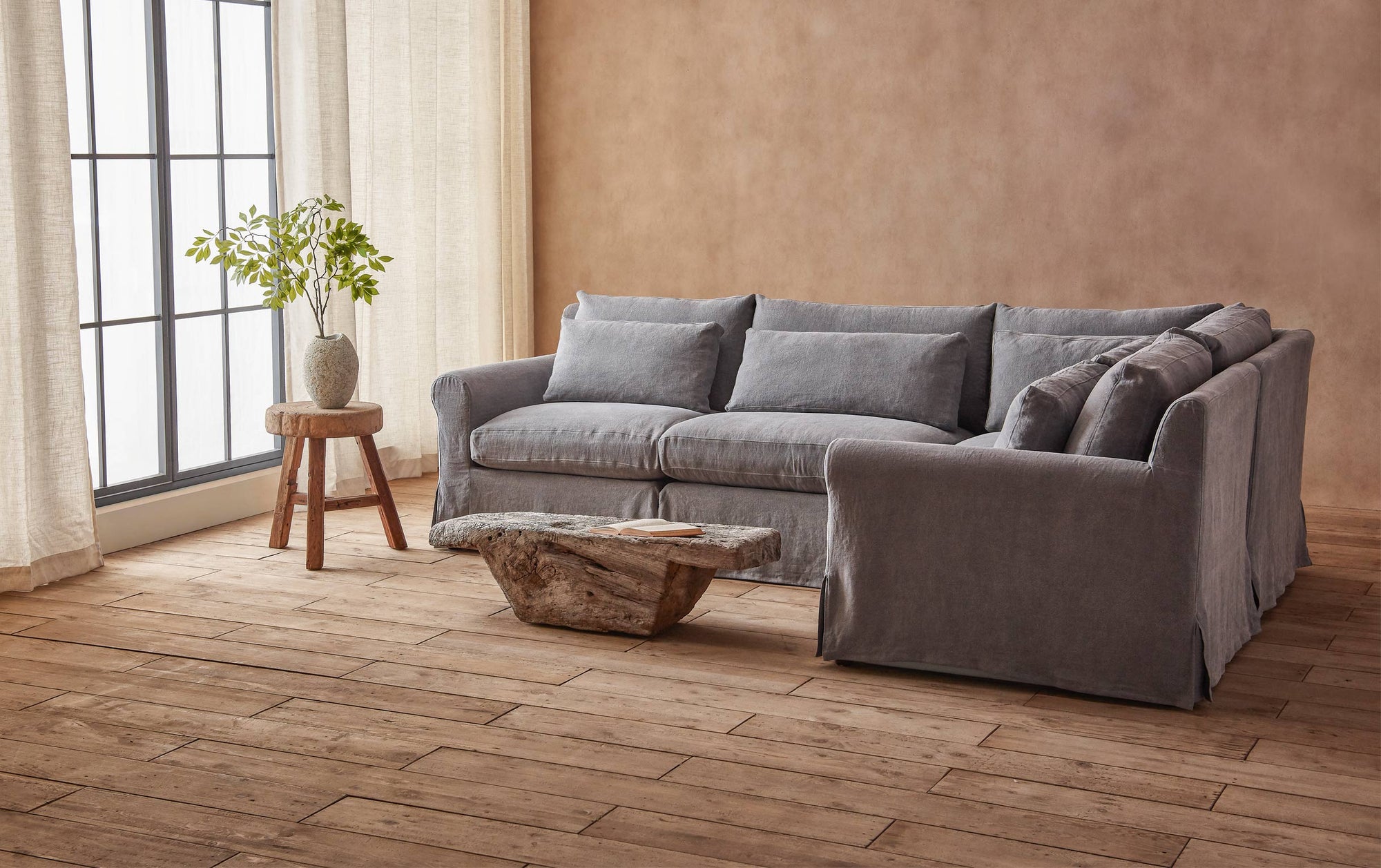 Elias L-Shape Sectional Sofa in Ink Cap, a medium cool grey Light Weight Linen, placed in a sunlit room next to a coffee table and a stool