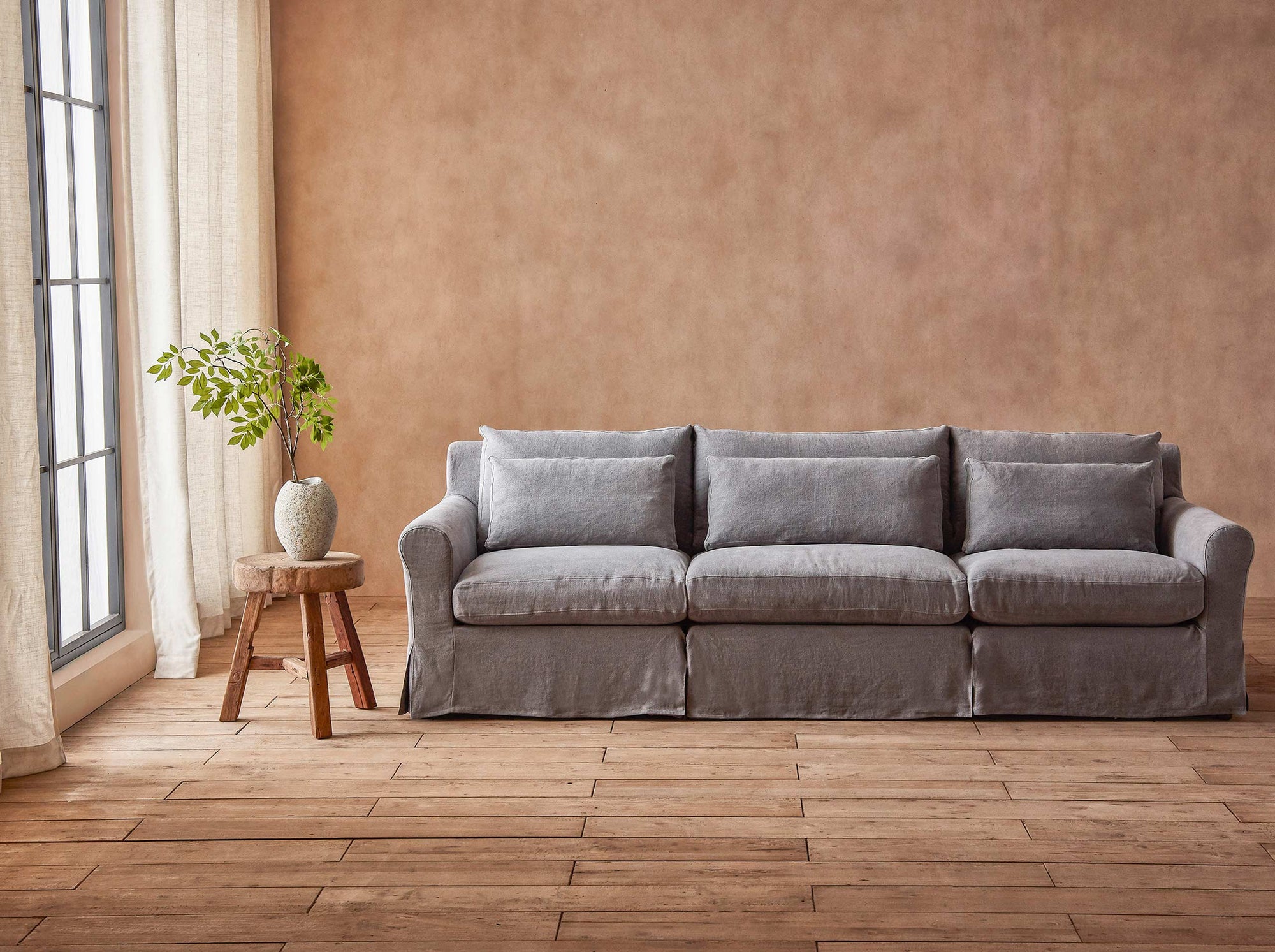 Elias Sectional Sofa in Ink Cap, a medium cool grey Light Weight Linen, placed in a sunlit room next to a plant in a vase on a stool