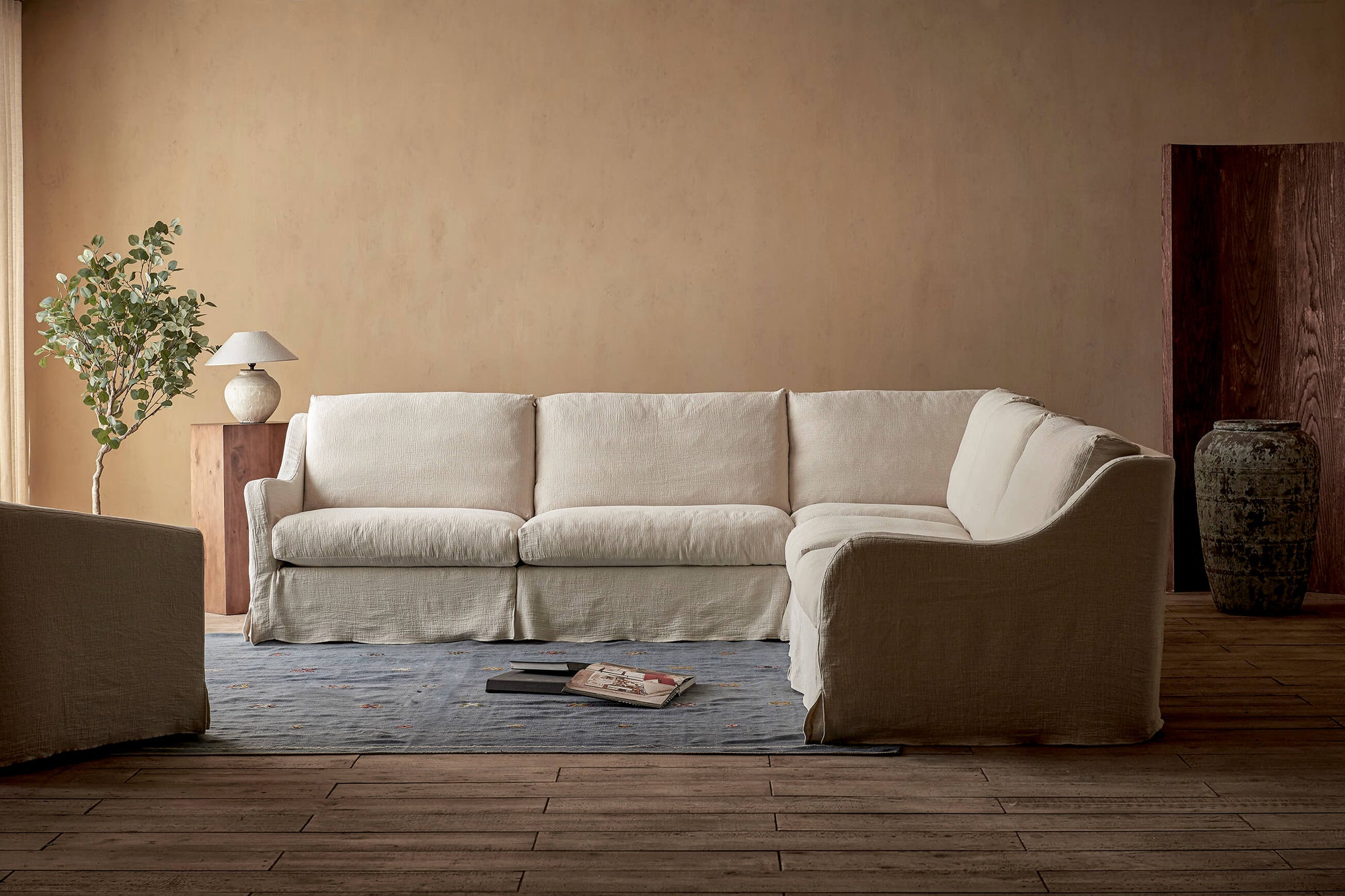 Esmé Corner Sectional Sofa in Corn Silk, a light beige Washed Cotton Linen, placed in a sunlit room decorated with a table lamp and a stack of books on the floor