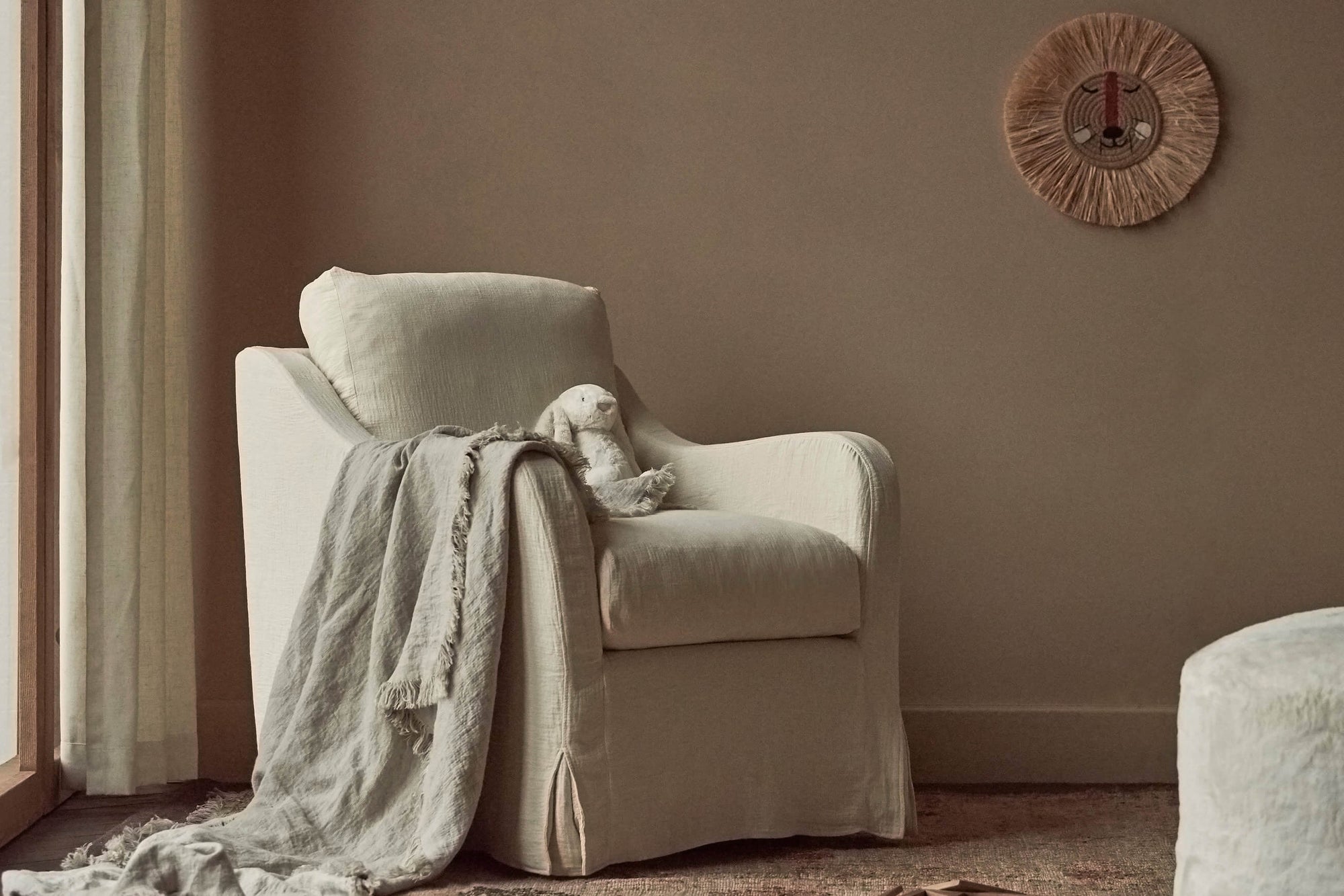 Esme Glider Chair in Corn Silk, a light beige Washed Cotton Linen, in a sunlit room by a window with a blanket and stuffed toy placed on the seat