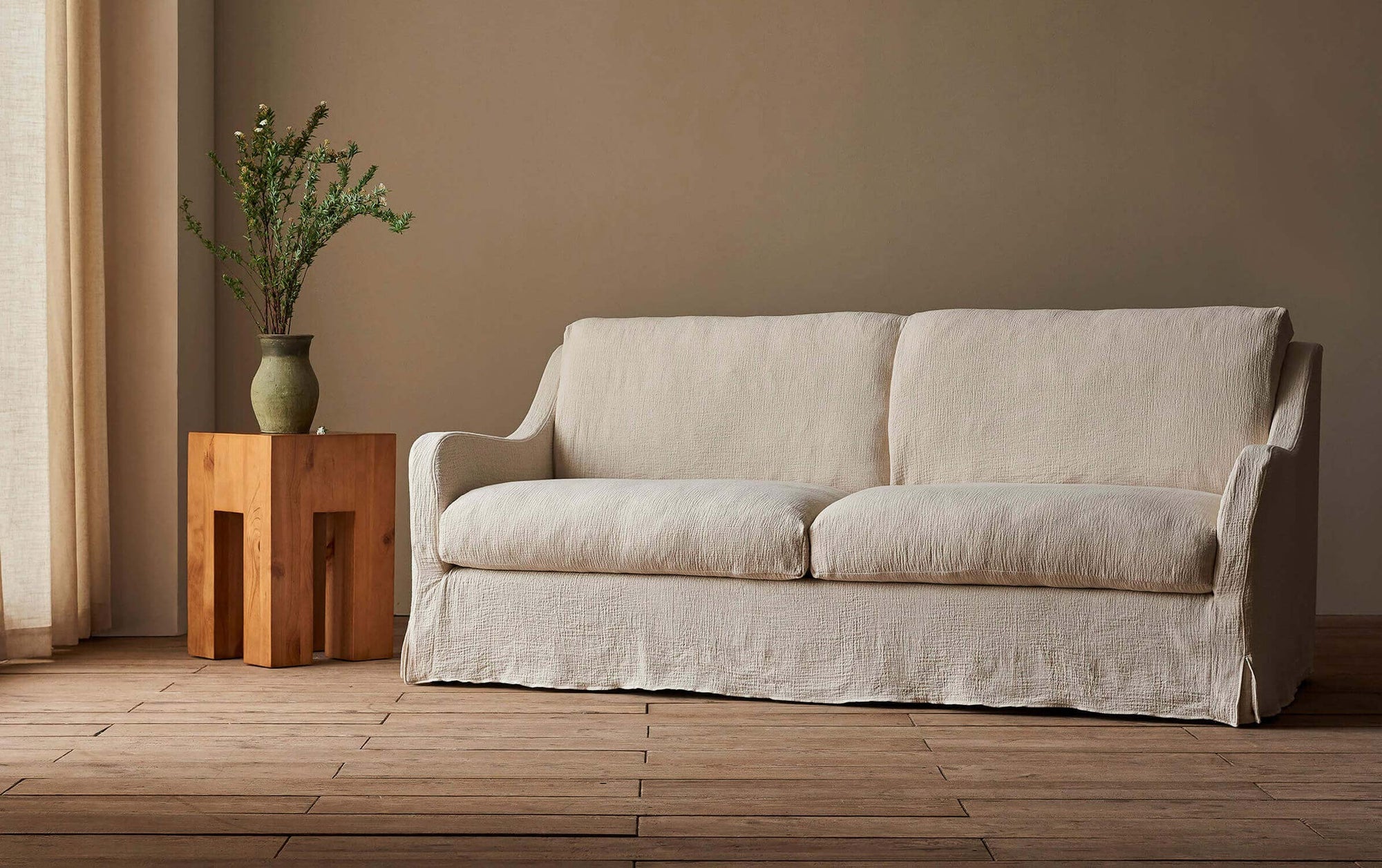 Esmé 84" Sofa in Jasmine Rice, a light warm greige Medium Weight Linen, next to a wooden side table decorated with a plant