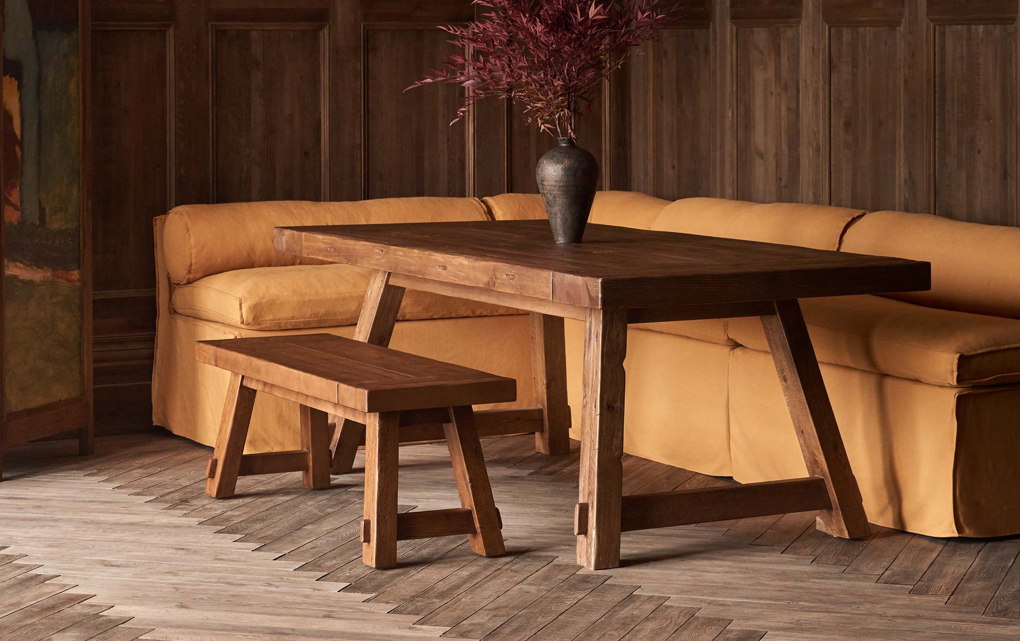 Fasso Dining Table in 100% Reclaimed Chinese Heritage Pine, a medium, bosc pear shade with earthy undertones, placed in a wood paneled room, between a Fasso Dining Bench and a Camino Banquette