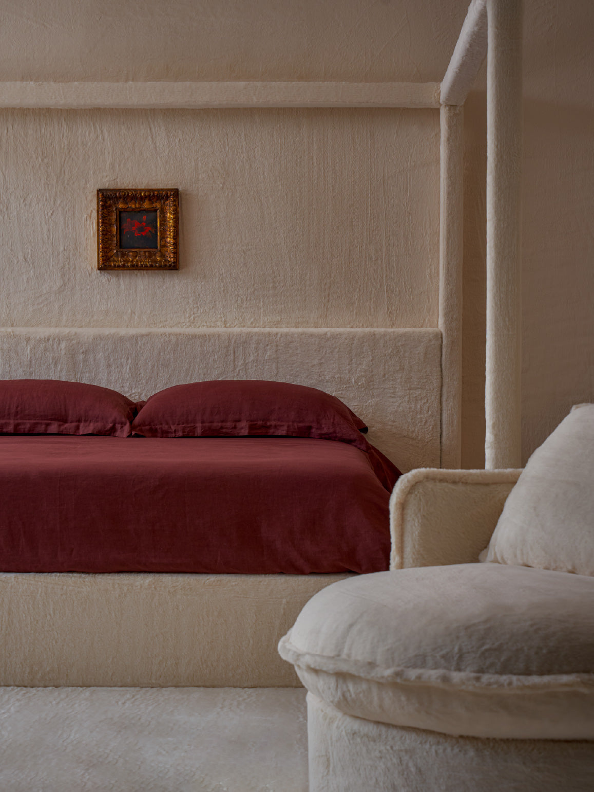 A minimalist bedroom scene featuring a florette bed in dandelion poof
neva round daybed in dandelion poof, set against a textured cream-colored wall and a matching upholstered headboard. A small framed artwork hangs on the wall, and a matching cream armchair is partially visible in the foreground.