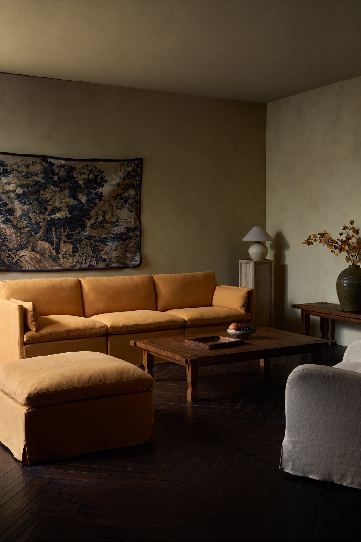 A warm, moody living room features a Sixpenny Gabriel Chaise Sectional in Thread-Dyed Cotton Linen in Honey Bee, paired with a matching ottoman, a dark wood coffee table, a textured wall tapestry, and dark herringbone wood flooring.