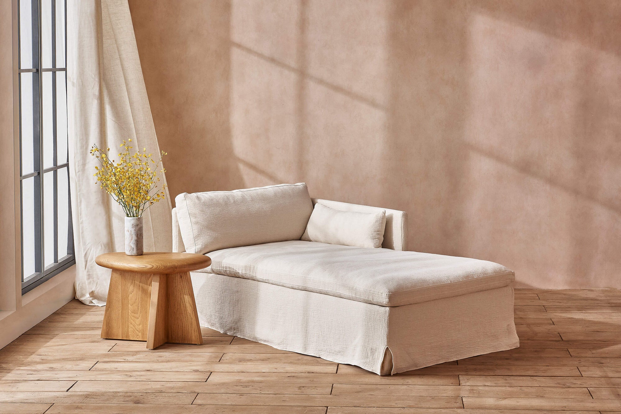 Gabriel Right Arm Facing Daybed in Corn Silk, a light beige Washed Cotton Linen, placed in a sunlit room next to a vase of flowers on a wooden end table