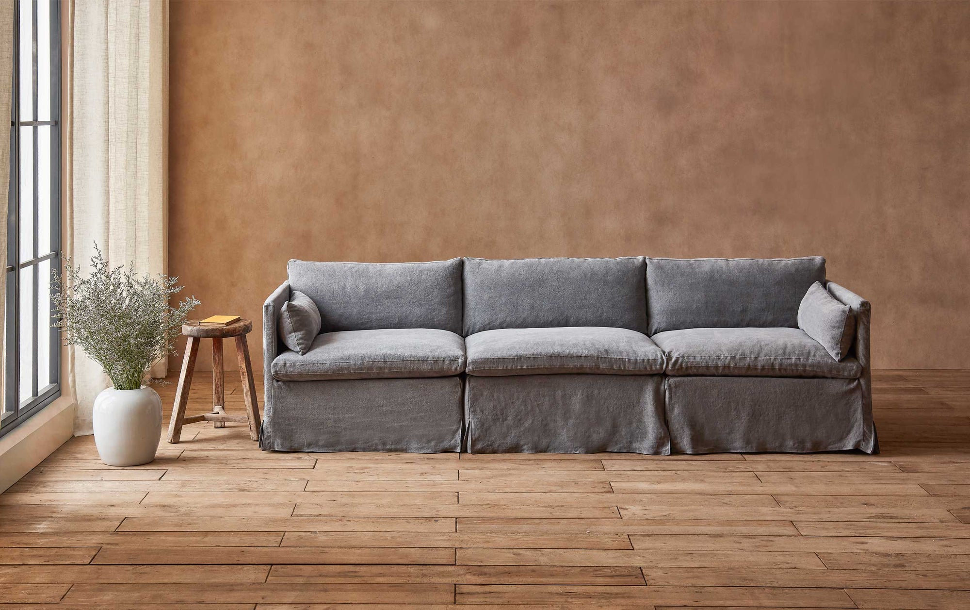 Gabriel Sectional Sofa in Ink Cap, a medium cool grey Light Weight Linen, placed in a sunlit room next to a stool