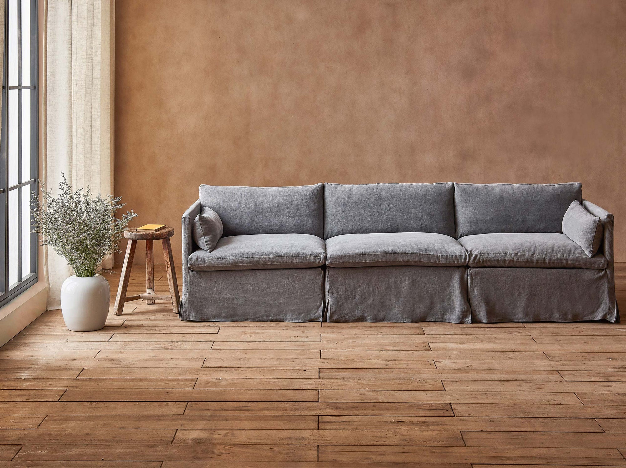 Gabriel Sectional Sofa in Ink Cap, a medium cool grey Light Weight Linen, placed in a sunlit room next to a stool