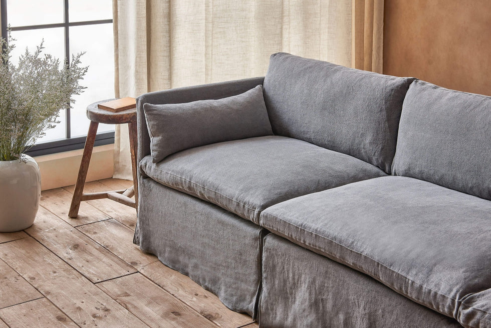 Close up of Gabriel Sectional Sofa in Ink Cap, a medium cool grey Light Weight Linen, placed in a sunlit room next to a stool and a vase of wildflowers